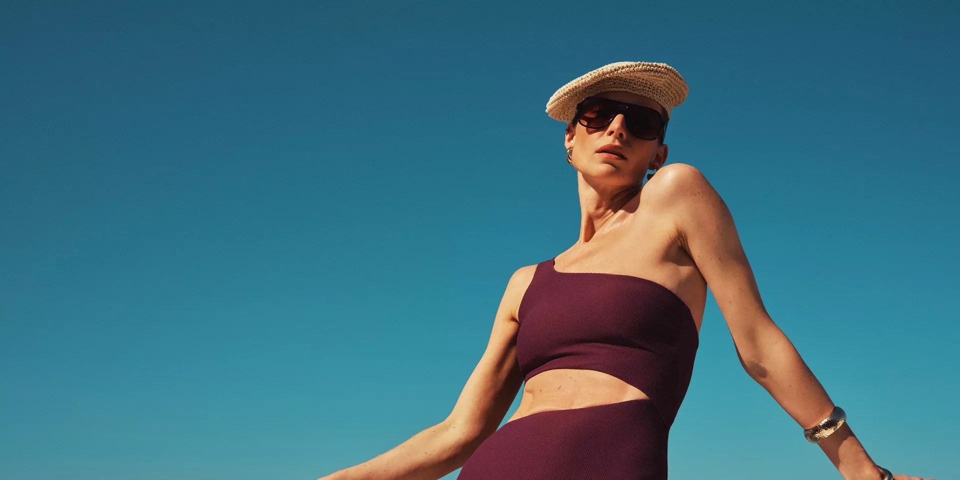 Woman in burgundy one-shoulder swimsuit and straw hat posing by sea rail under clear blue sky