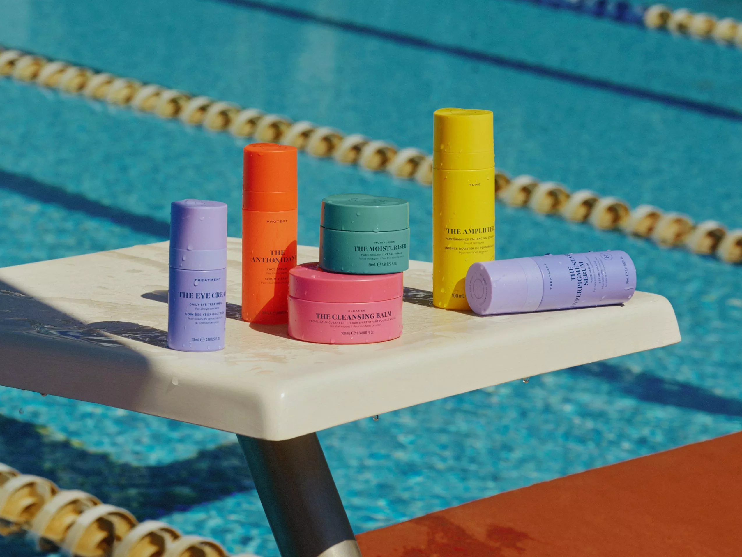 Colourful skincare products on a poolside diving board, including moisturiser, eye cream, serum and cleansing balm.