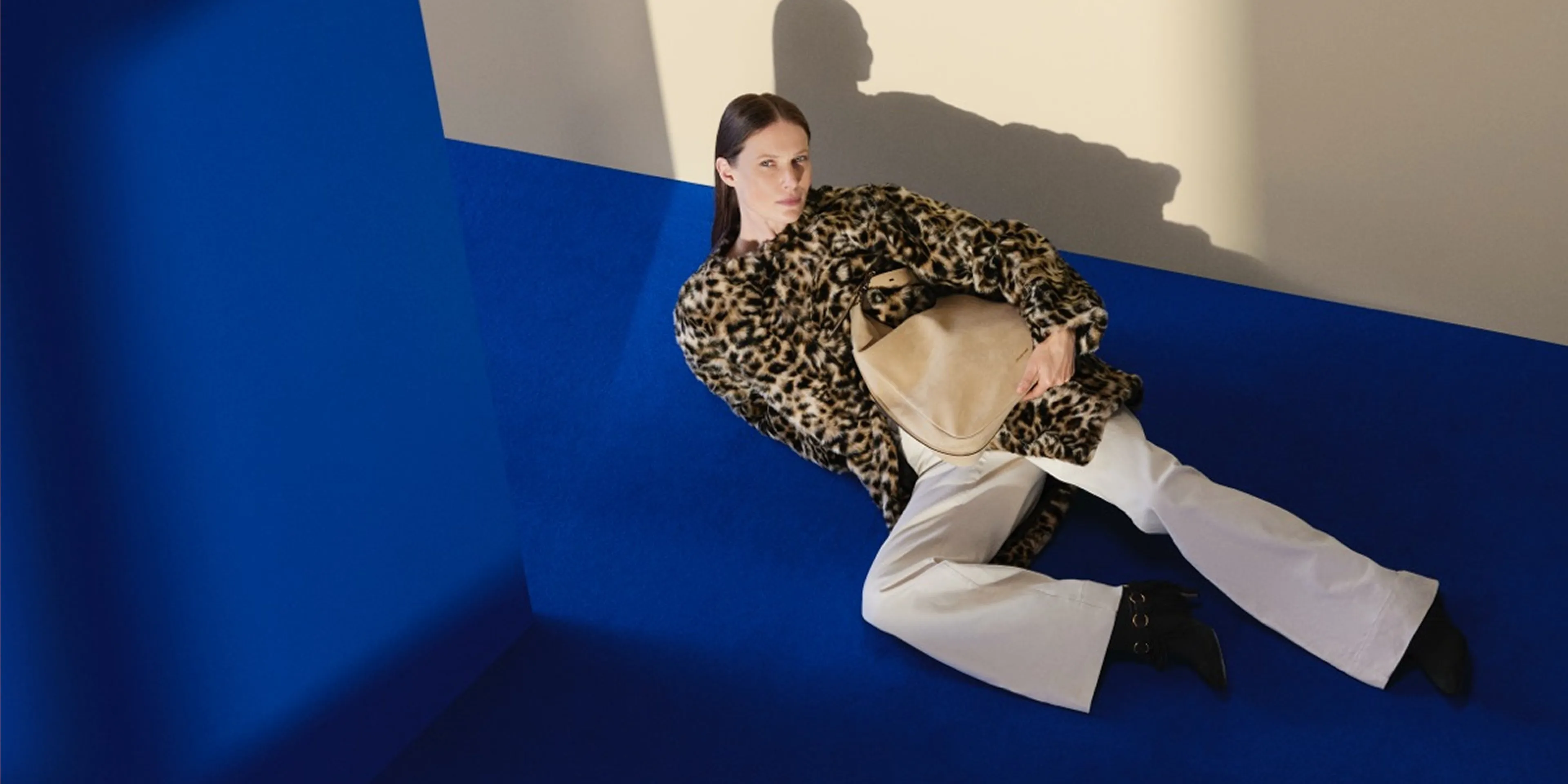 Fashion model in leopard-print coat holding beige handbag, reclining on blue studio floor in dramatic light.