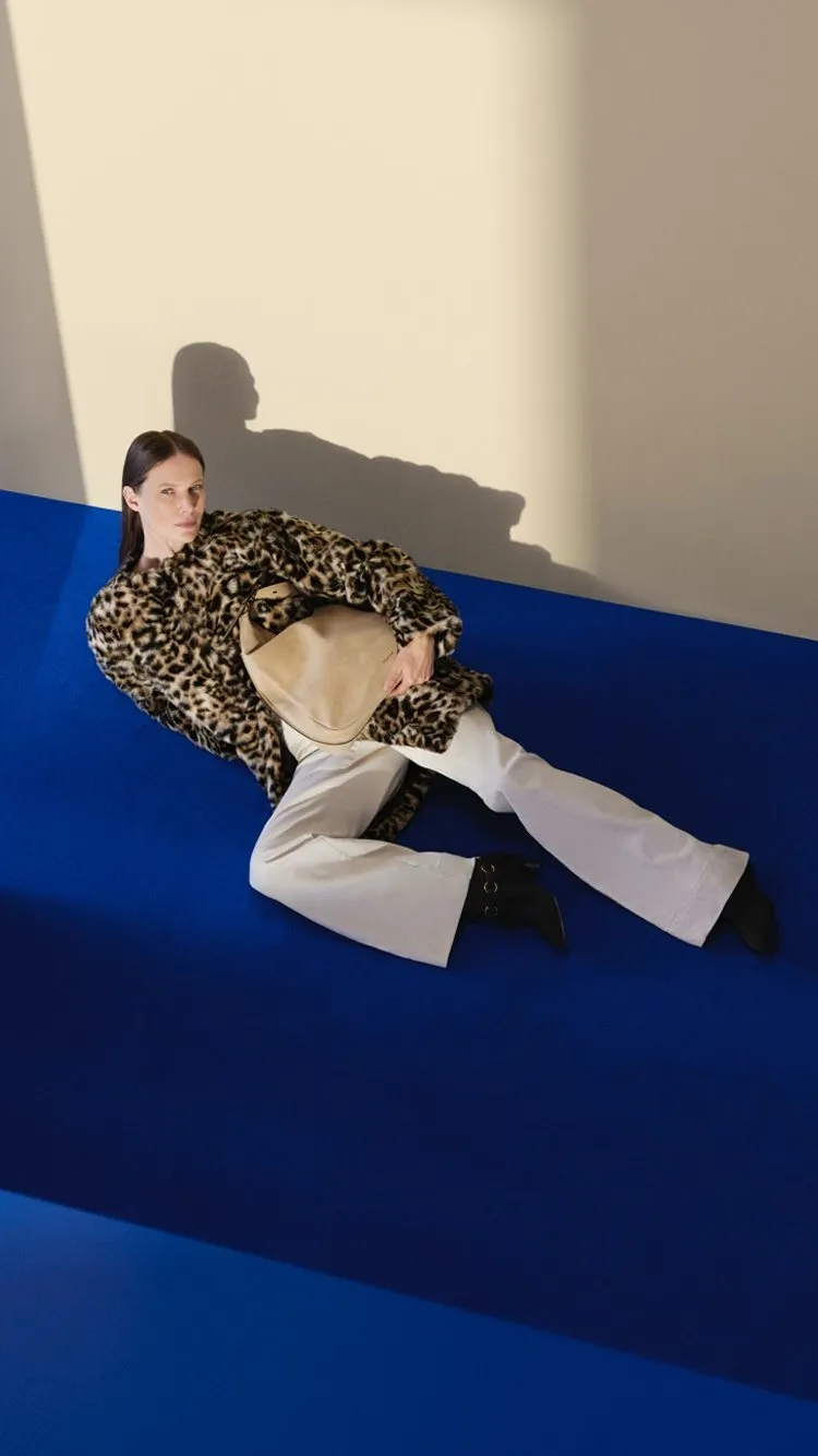 Fashion model in leopard-print coat holding beige handbag, reclining on blue studio floor in dramatic light.