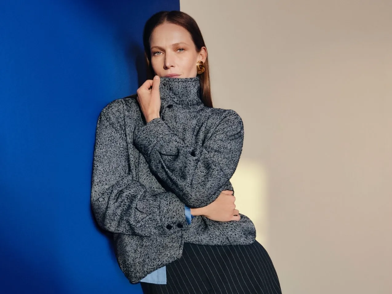 Fashion portrait of woman in grey tweed high-neck jacket and pinstripe skirt against bold blue wall backdrop