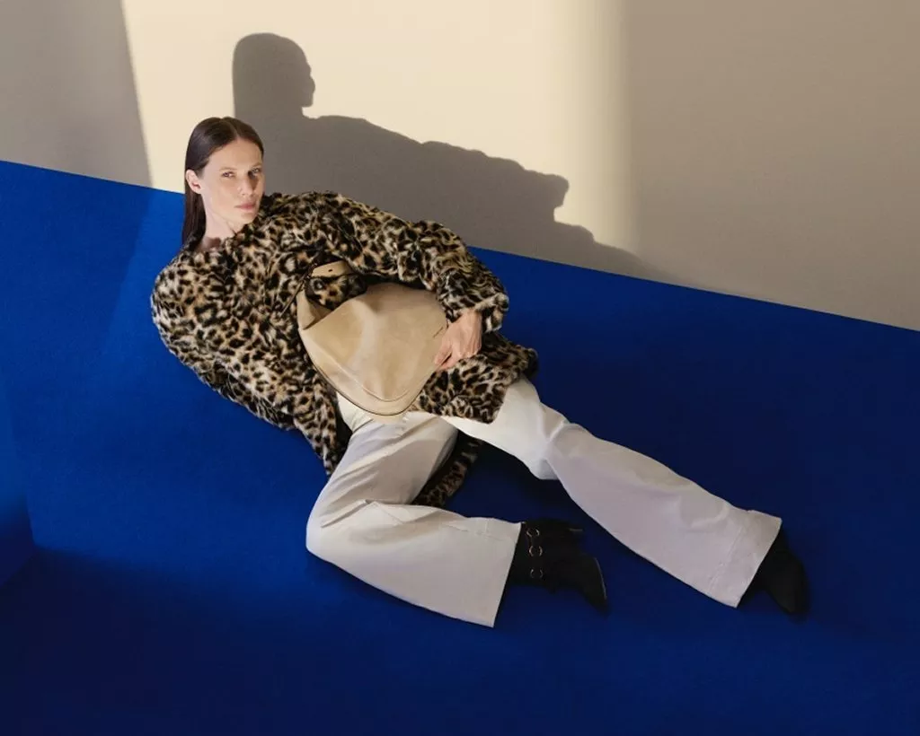 Fashion model reclining on blue floor in leopard print coat and wide-leg trousers, holding beige handbag