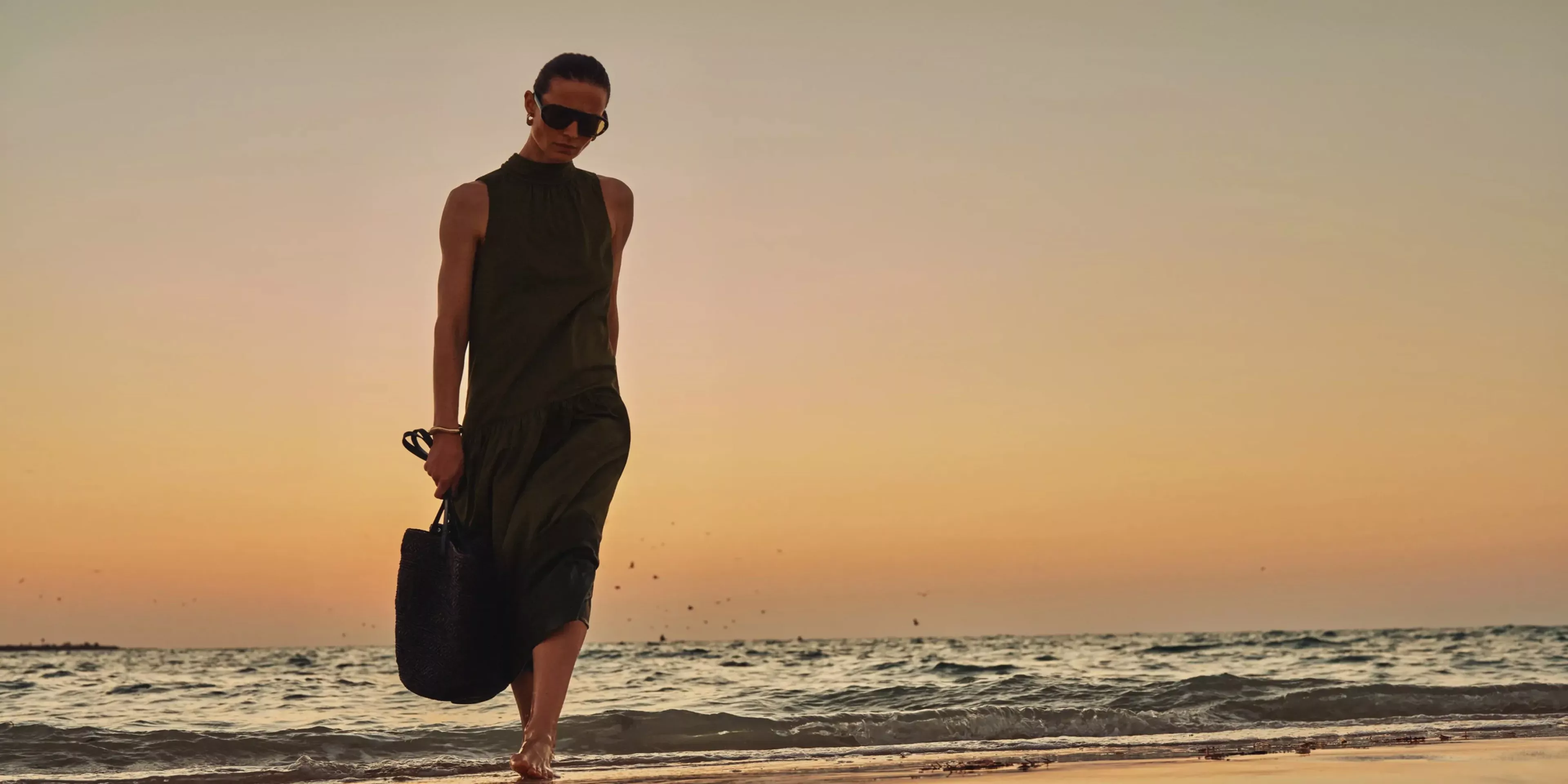 Fashion model in dark green sleeveless dress and sunglasses walking barefoot on a beach at sunset, holding bag