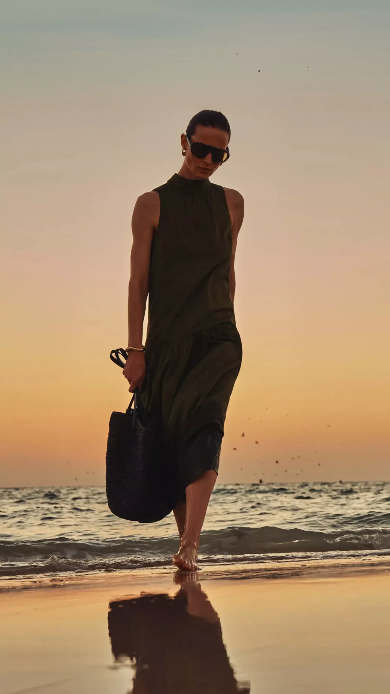 Fashion model in dark green sleeveless dress and sunglasses walking barefoot on a beach at sunset, holding bag