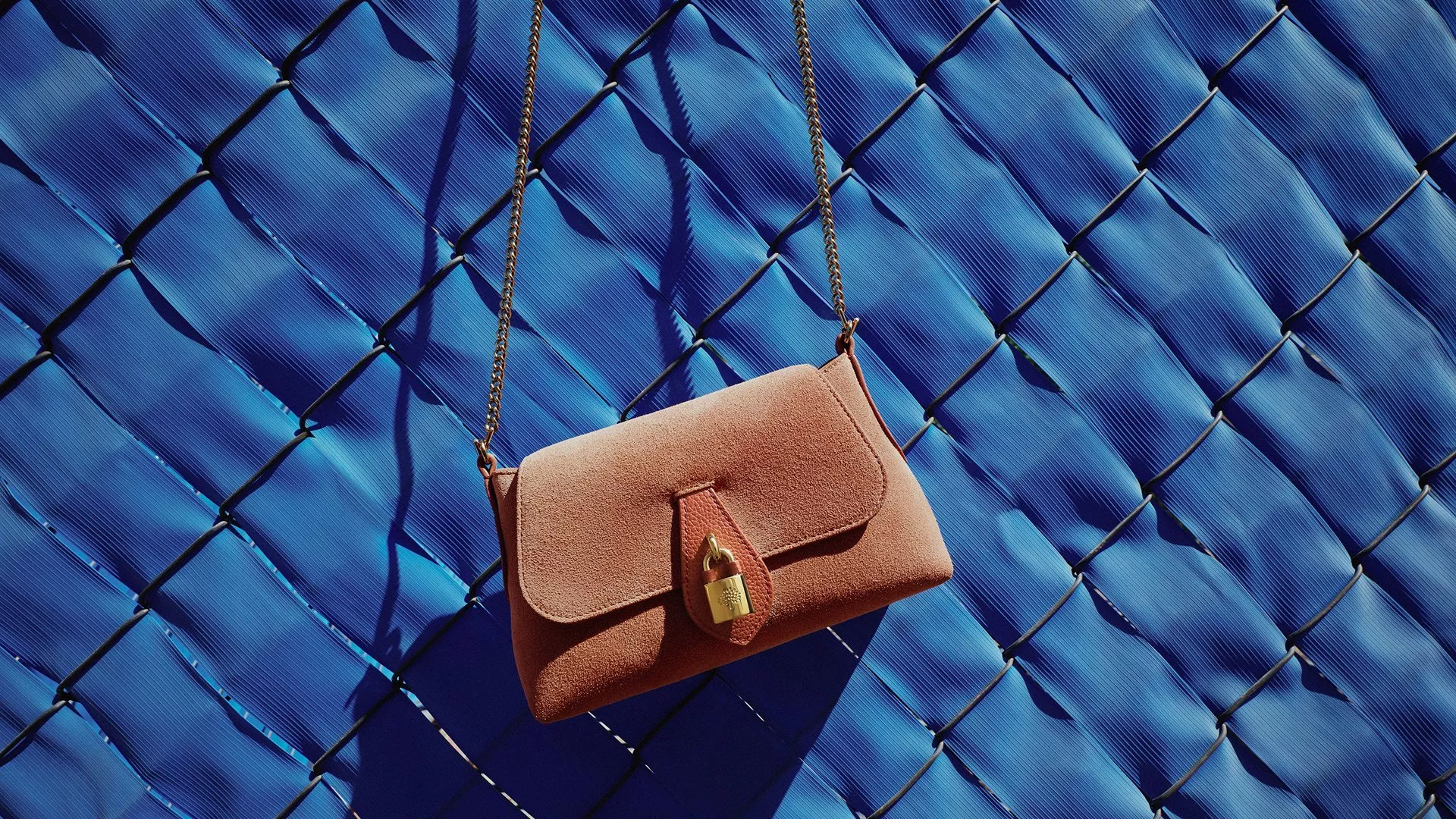 Pink suede crossbody handbag with gold chain strap on blue fence background