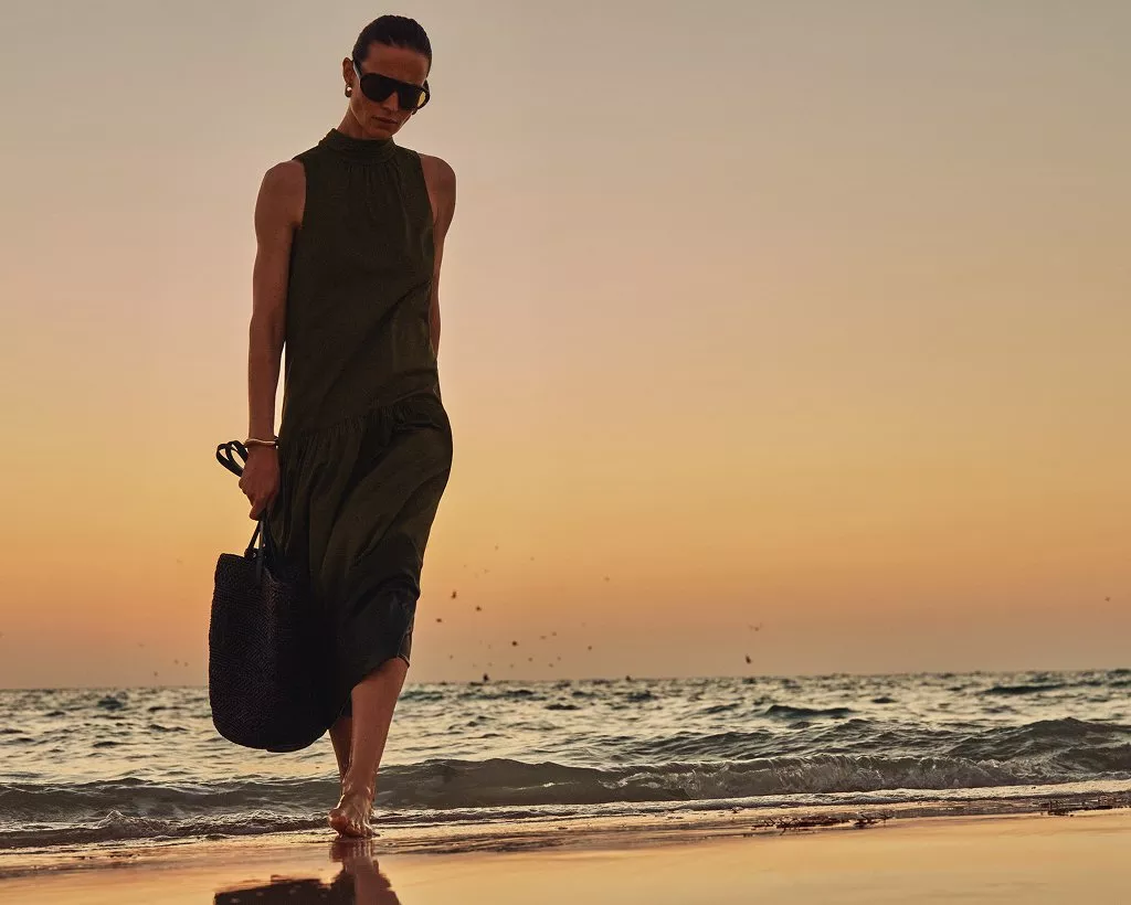Fashion model in sleeveless green dress and sunglasses walking barefoot on beach at sunset, holding woven bag