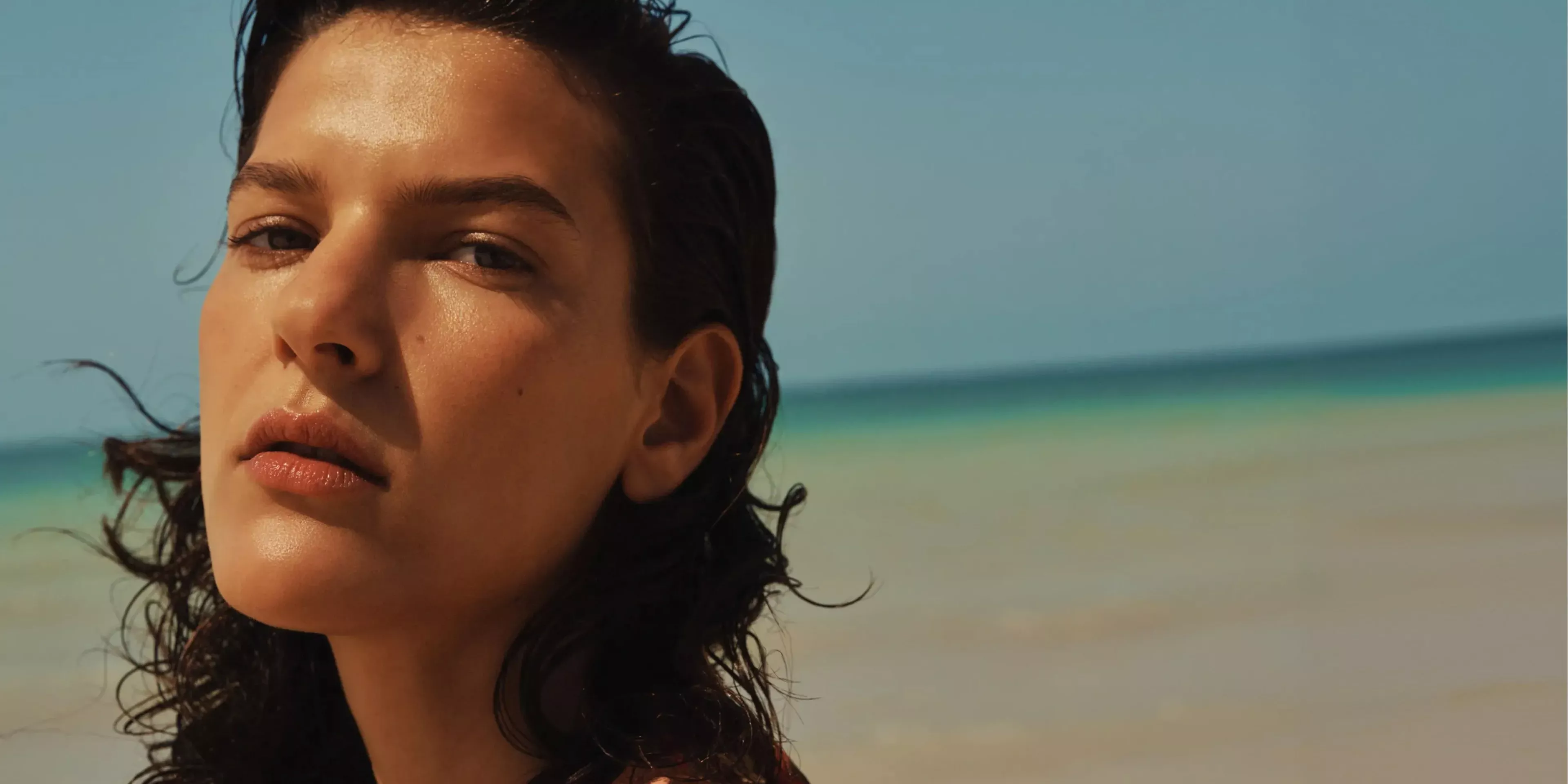 Close-up portrait of a woman with wet dark hair on a sunny beach, turquoise sea behind, fashion editorial style