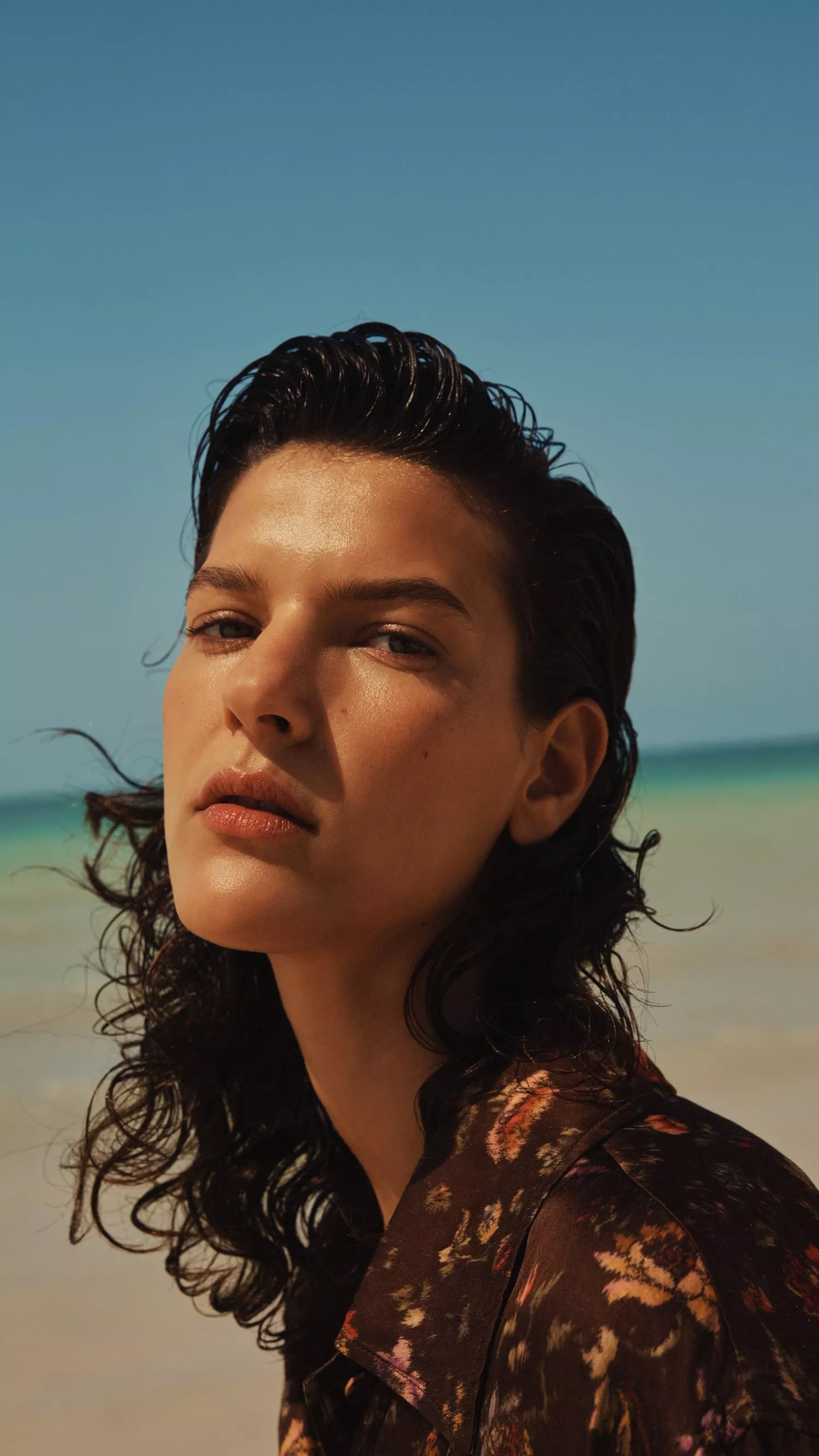 Close-up portrait of a woman with wet dark hair on a sunny beach, turquoise sea behind, fashion editorial style