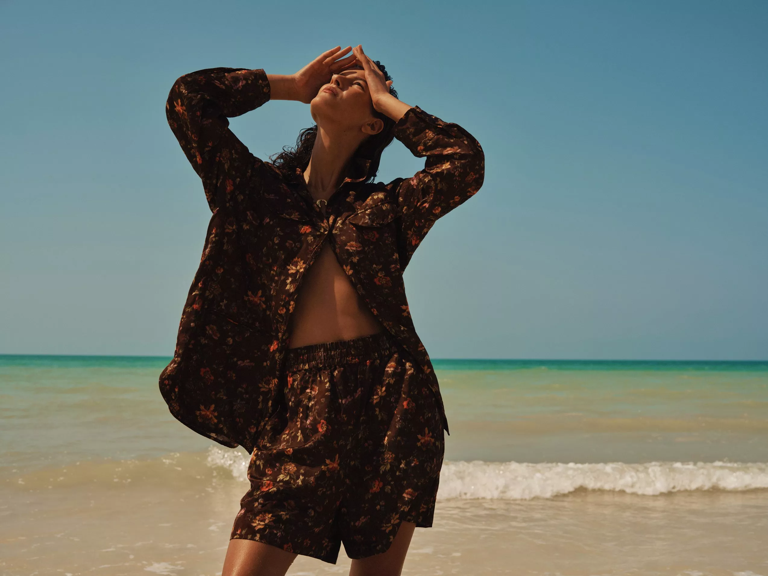 Woman posing on a sunny beach in a floral co-ord set, hands shielding eyes, with sea and blue sky behind