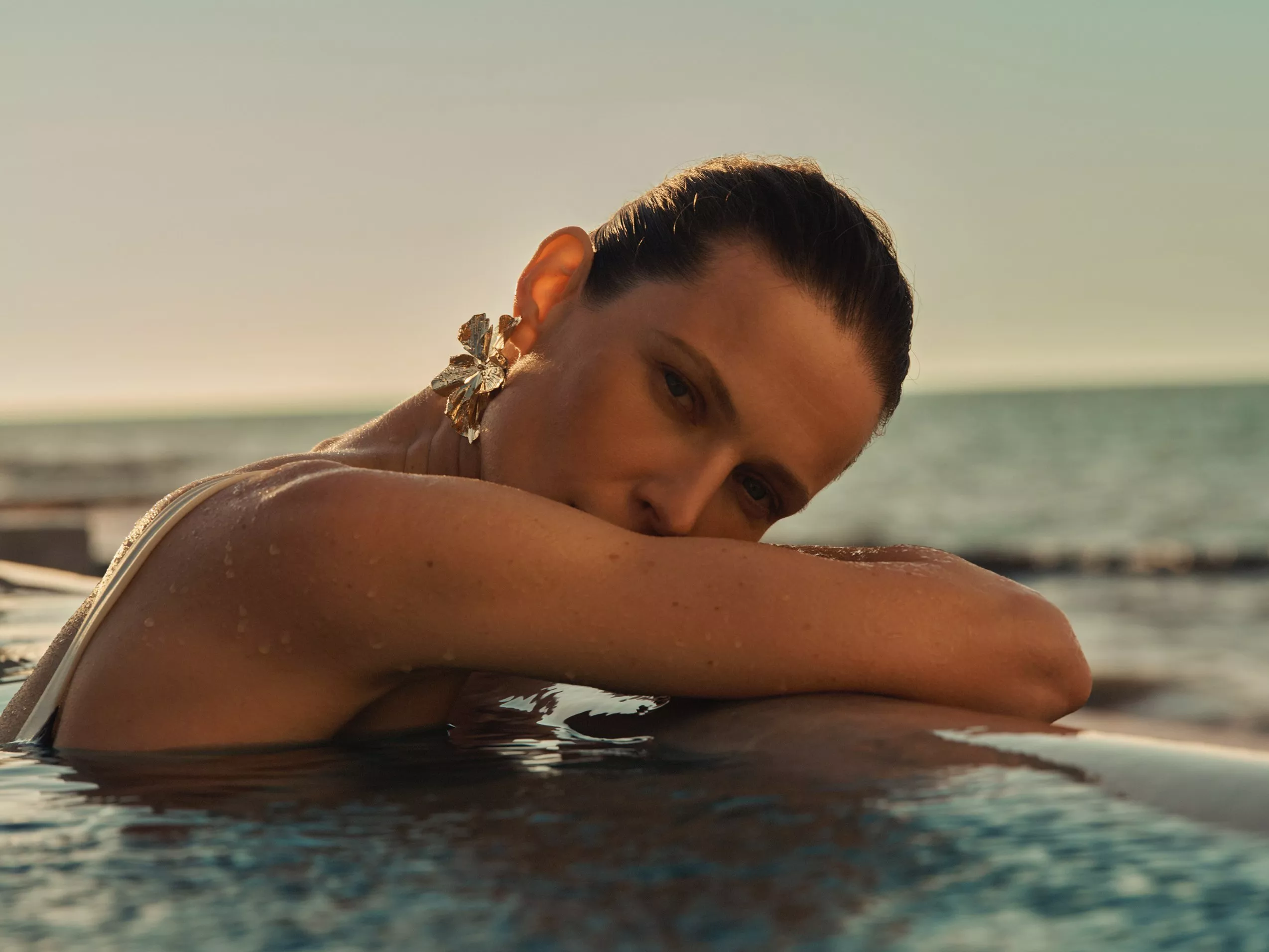 Close-up of woman in infinity pool at sunset, wearing statement floral earrings with sea horizon backdrop