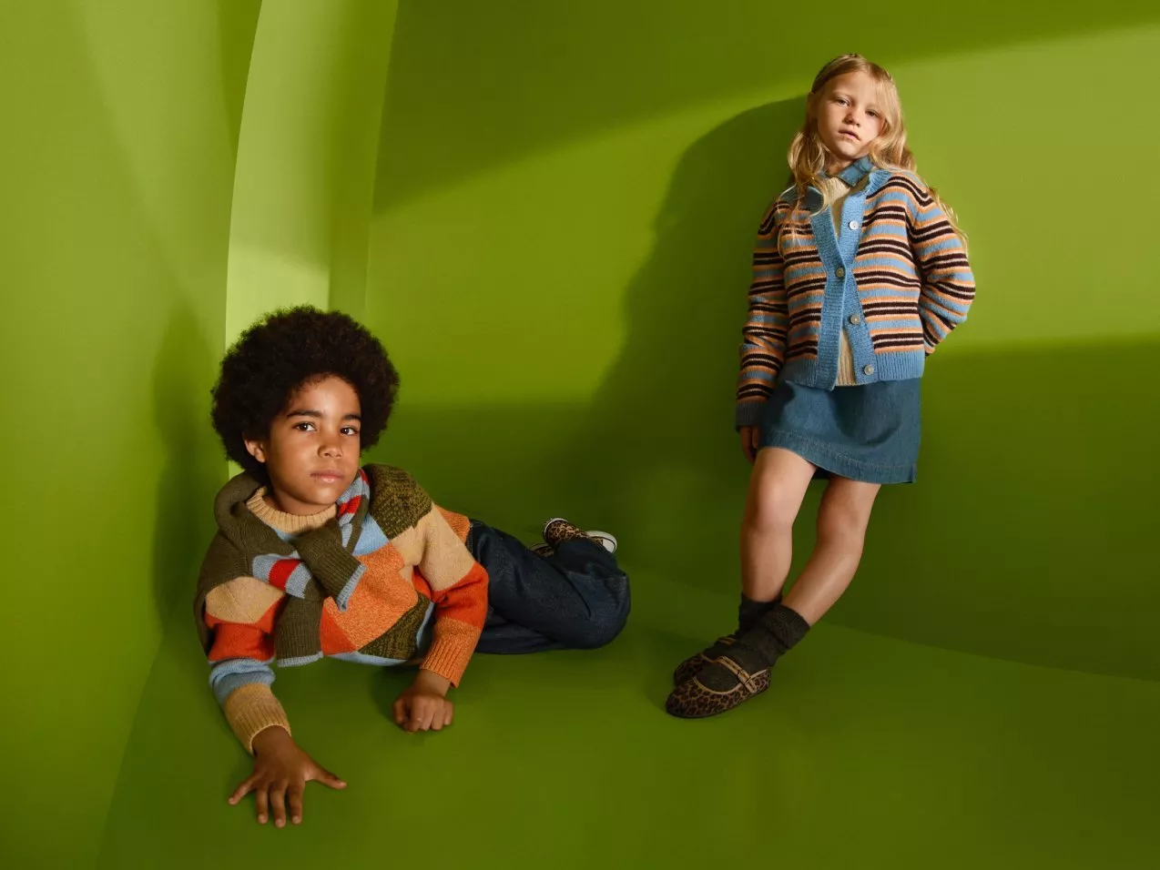 Two children modelling colourful knitwear and denim in a green studio, stylish autumn fashion portrait.