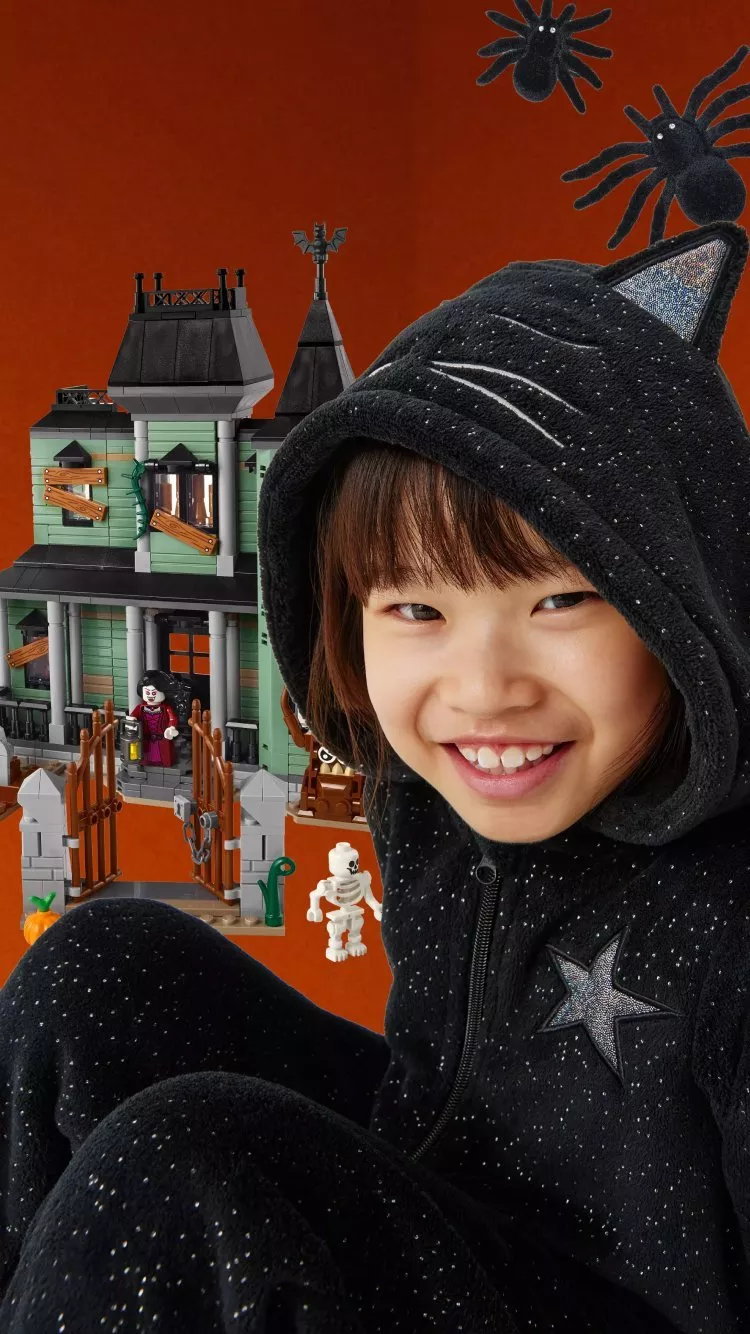 Smiling child in black cat hoodie by spooky LEGO haunted house set with bats, spiders and skeleton, Halloween playtime