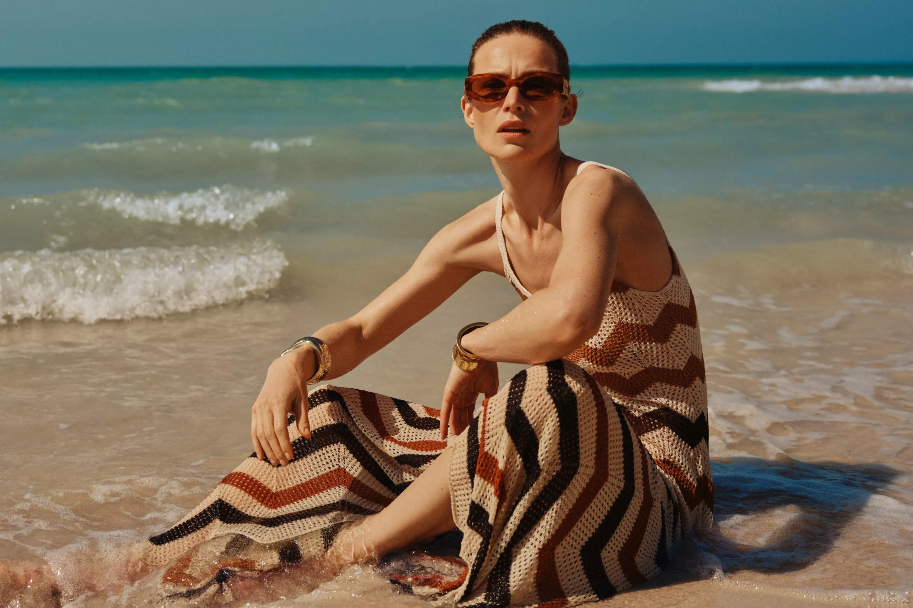 a women in a sundress sitting on the beach with water lapping around her