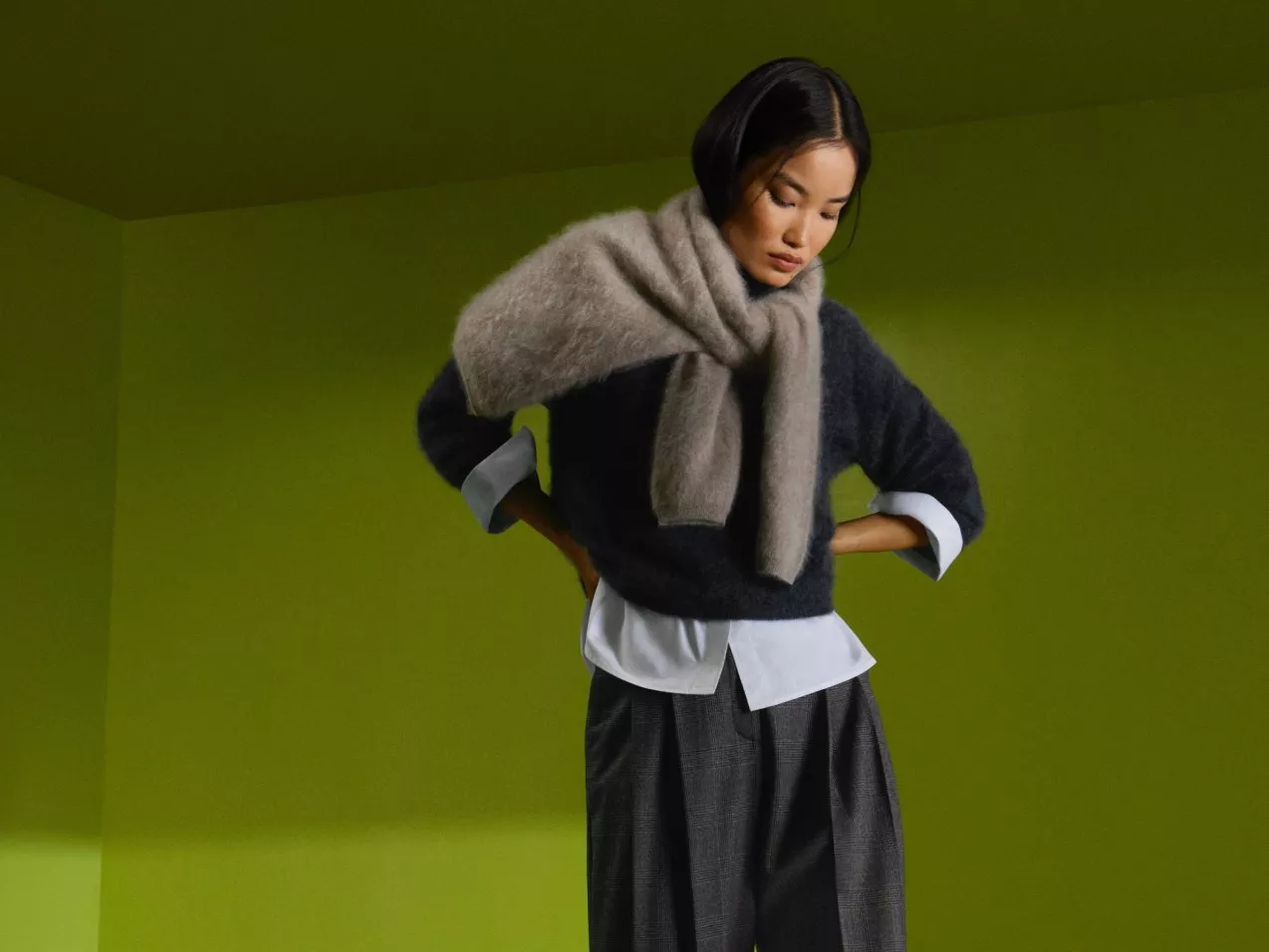 Fashion model in grey knit jumper and oversized scarf, layered over white shirt and trousers on green backdrop
