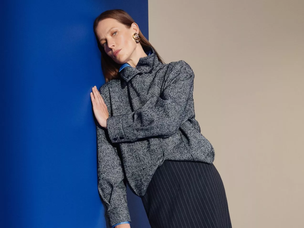 Fashion model in grey tweed jacket and pinstripe skirt leaning against blue wall, modern studio portrait