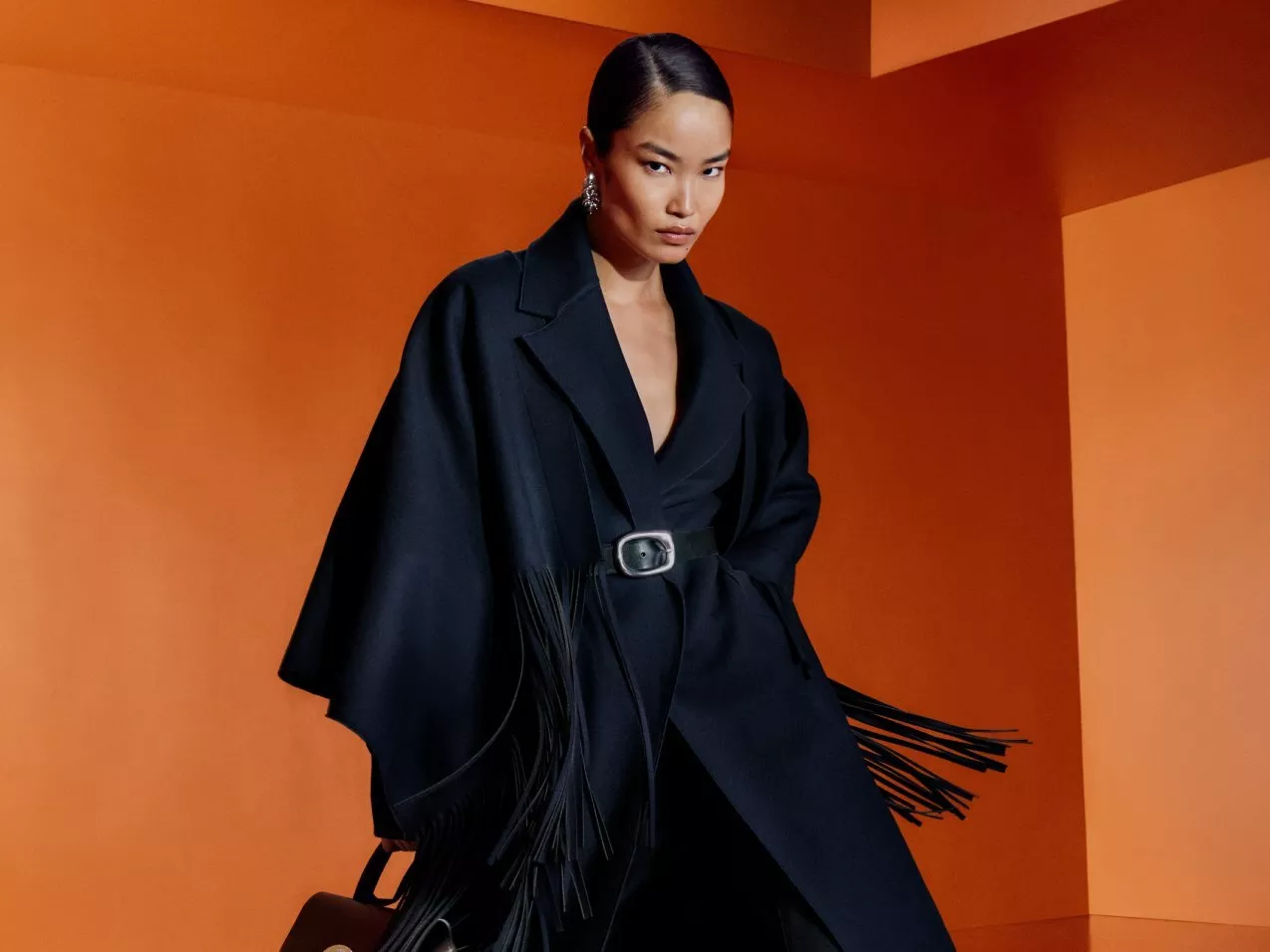 A woman in a belted black fringe coat against a bold orange studio backdrop, editorial style