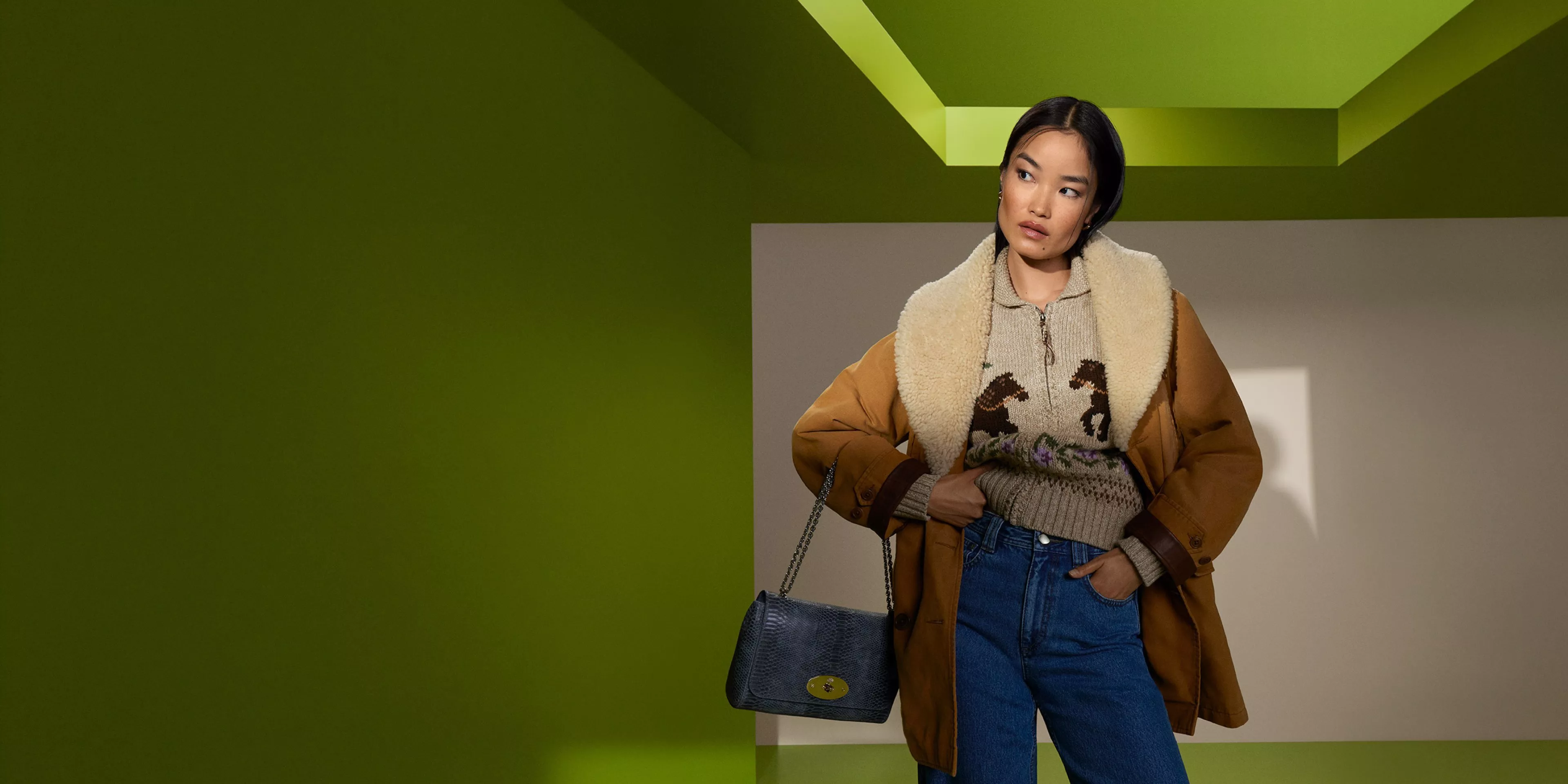 Woman in shearling-lined tan jacket, knit jumper and wide-leg jeans, holding blue handbag on green set
