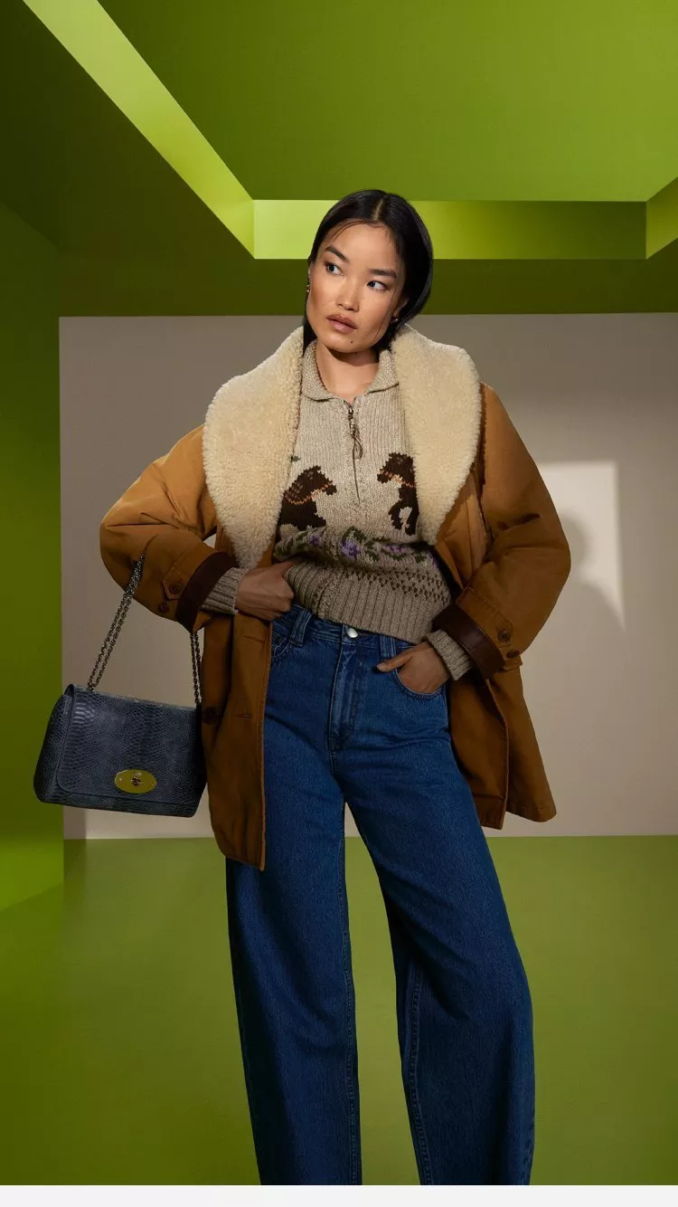 Woman in shearling-lined tan jacket, knit jumper and wide-leg jeans, holding blue handbag on green set