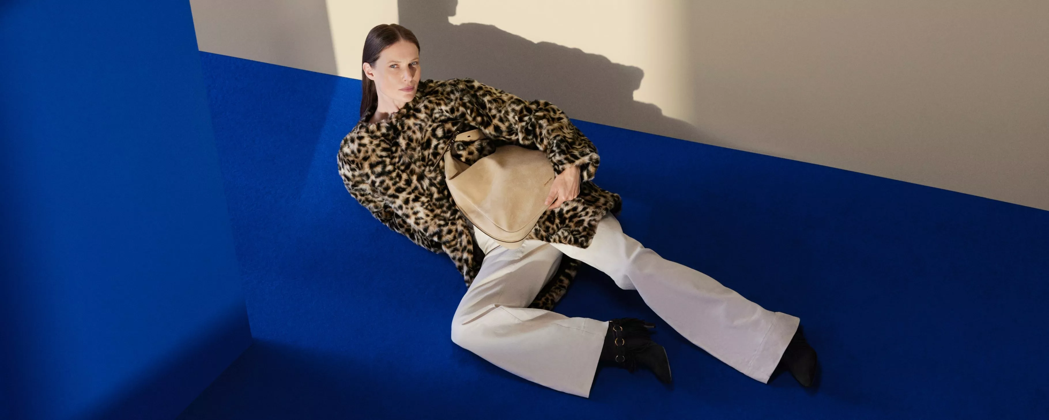 Woman in leopard print faux fur coat holding beige handbag, reclining on bold blue set background