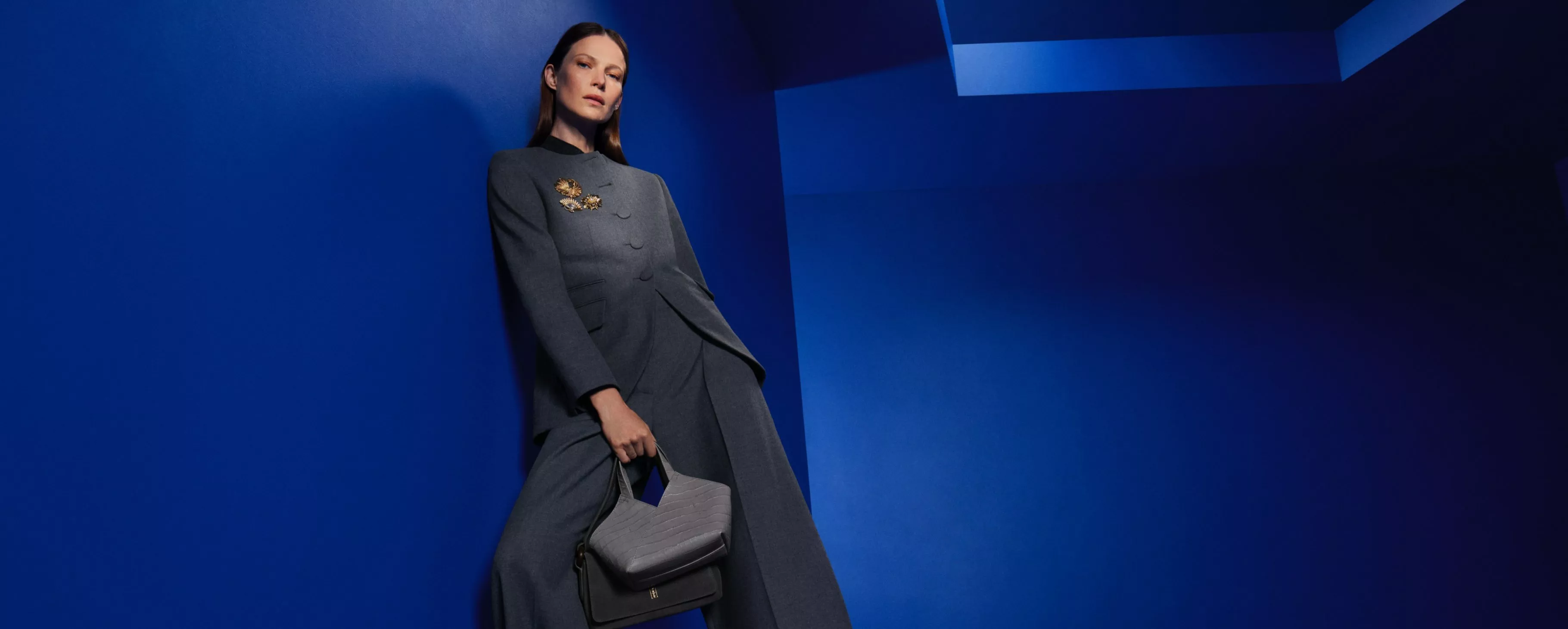 a woman in grey tailored suit holding structured handbag, posing against bold blue studio backdrop