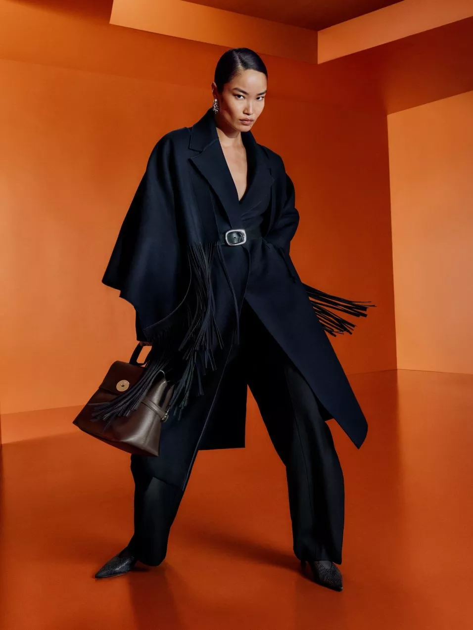 A woman in oversized black belted coat with fringed details and handbag, posing in orange studio set
