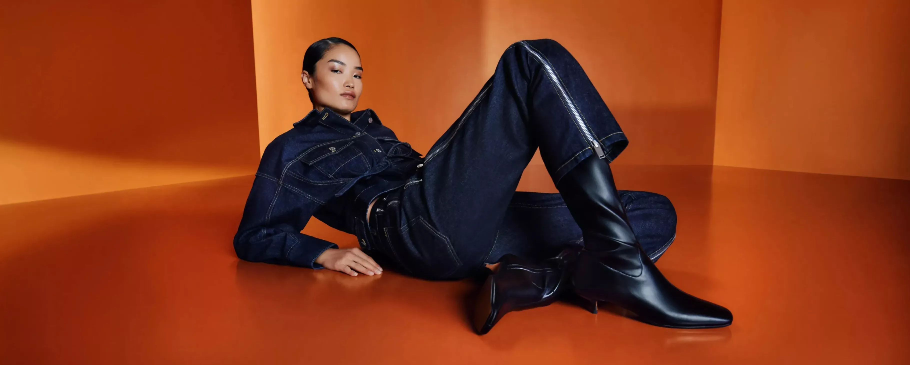 A woman reclining in dark denim jacket and jeans with black knee-high boots on orange backdrop
