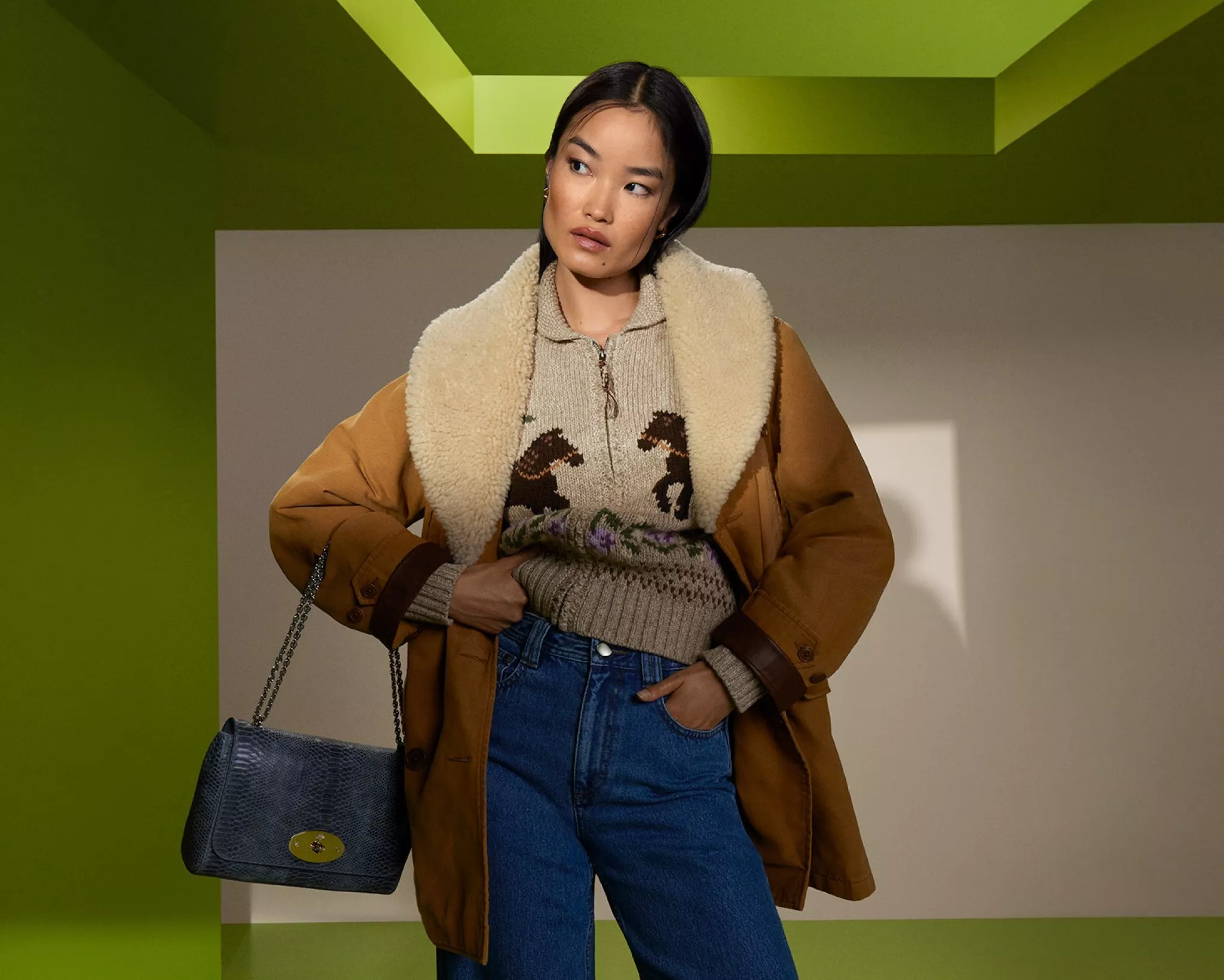 A woman in shearling-lined tan coat, knit jumper and jeans, holding chain strap handbag in studio setting