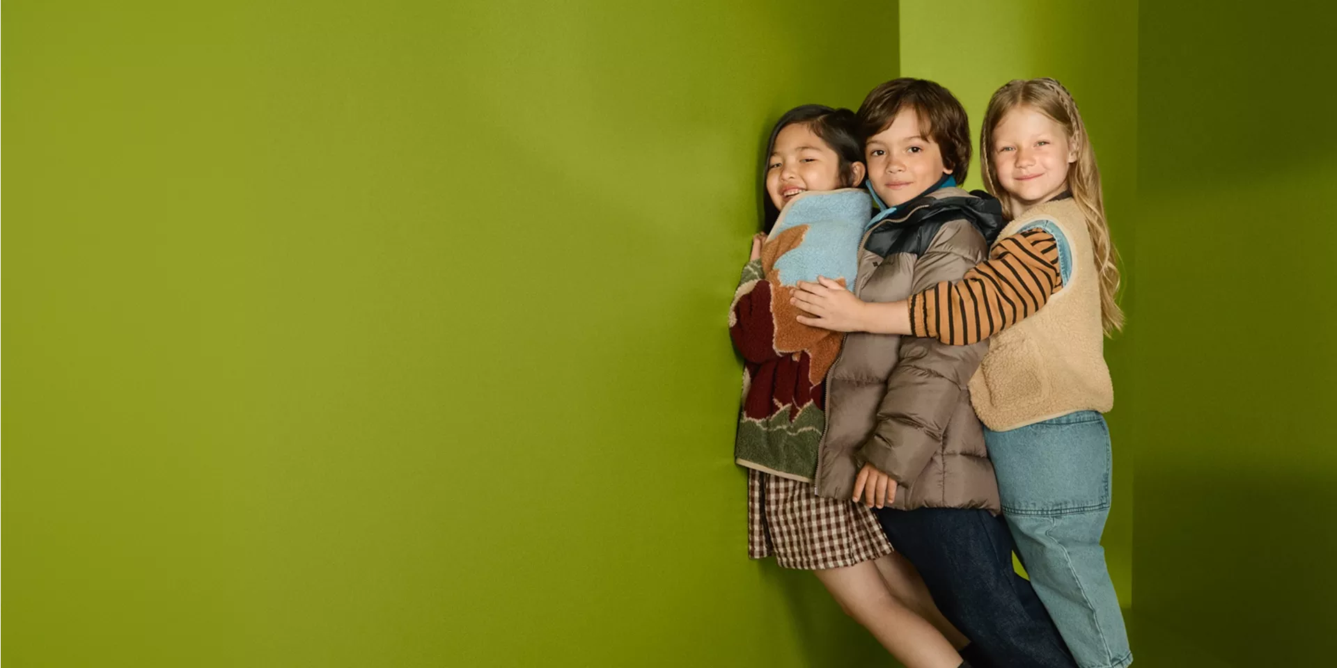 Three smiling children hugging in colourful autumn outfits against a bright green studio background