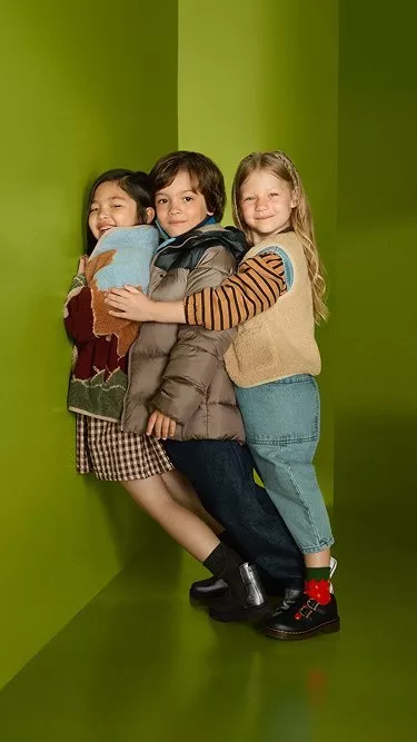 Three smiling children hugging in colourful autumn outfits against a bright green studio background