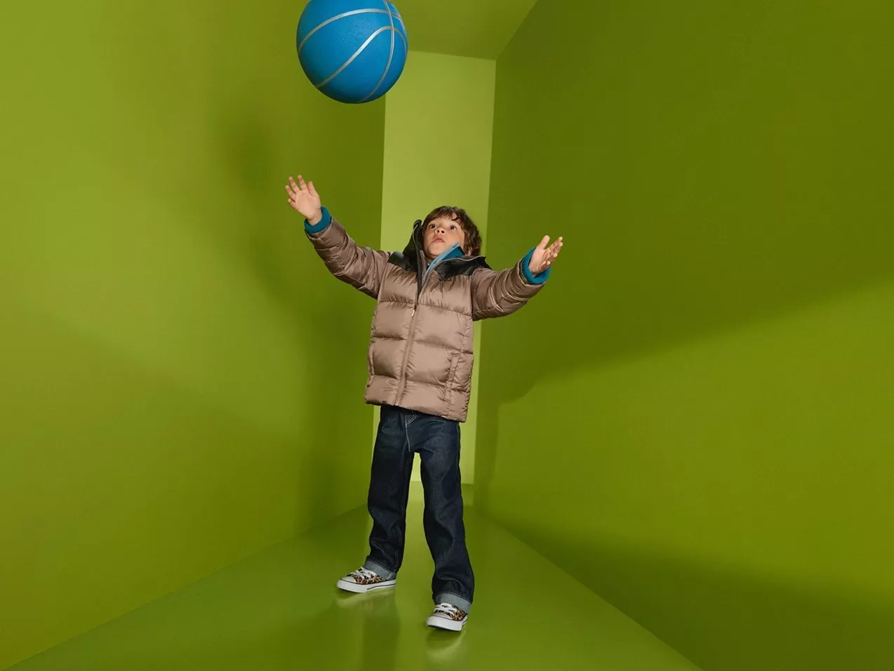 Child in winter coat tossing blue ball in bright green room, playful indoor kids activity and fun game