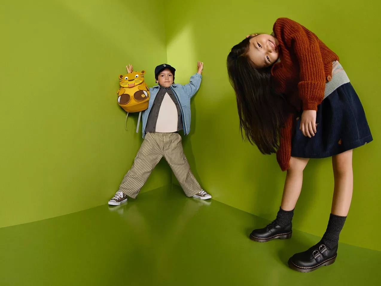 Two children in stylish autumn outfits posing in a green studio corner, showcasing kids fashion and footwear