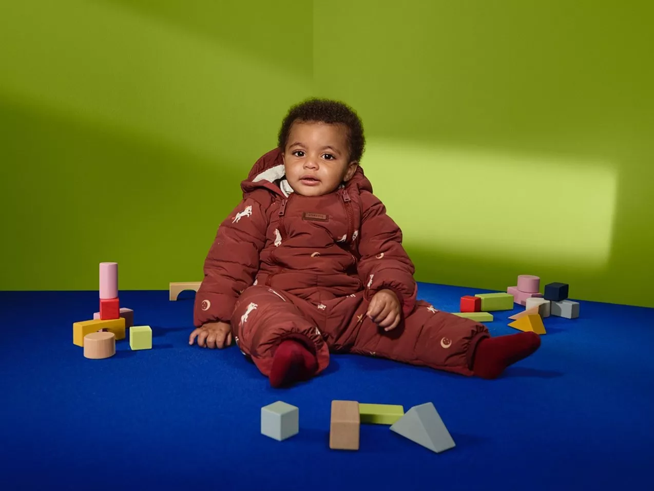 Baby in burgundy snowsuit sitting on blue mat with wooden blocks, green studio backdrop