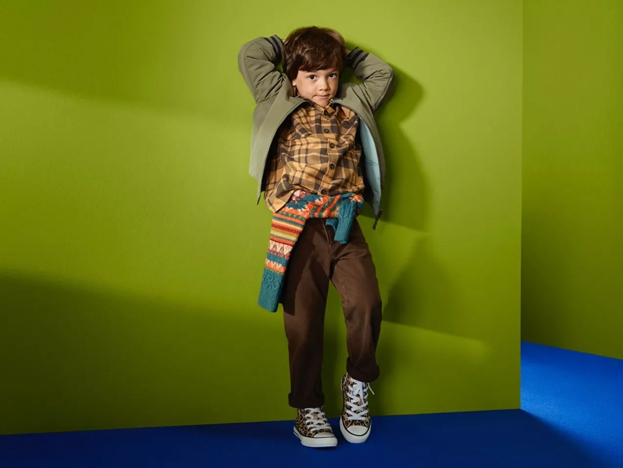 Fashionable young boy in plaid shirt, green jacket and patterned scarf, leaning against green wall in trainers