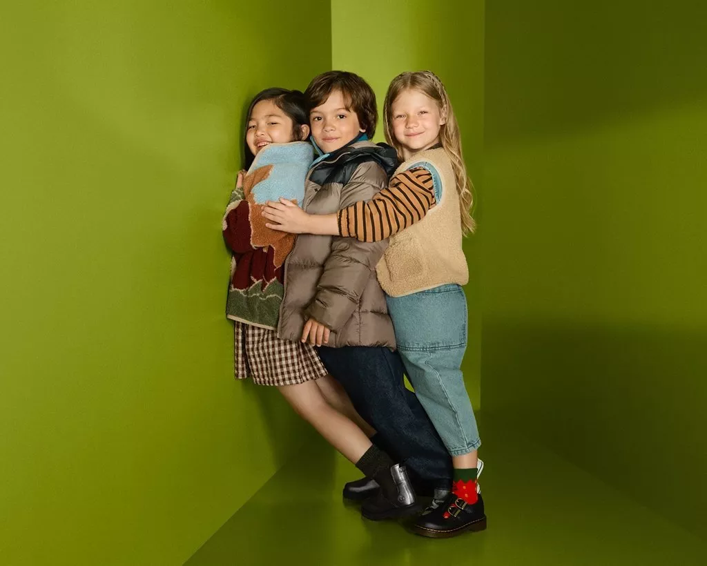 Three children hugging in green studio corner, wearing cosy autumn outfits, knitwear and puffer jacket, smiling.