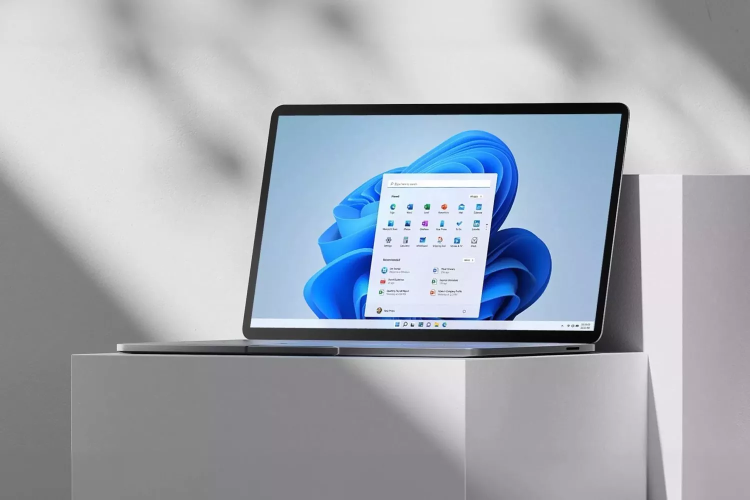 Laptop showing Windows 11 Start menu on screen, on minimalist desk with soft shadows and grey background