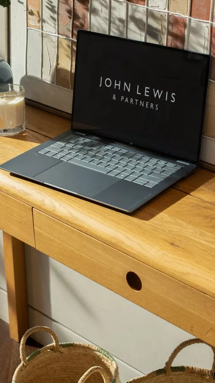 Laptop displaying John Lewis & Partners on a wooden desk by a tiled wall, with candle and sunlight shadows