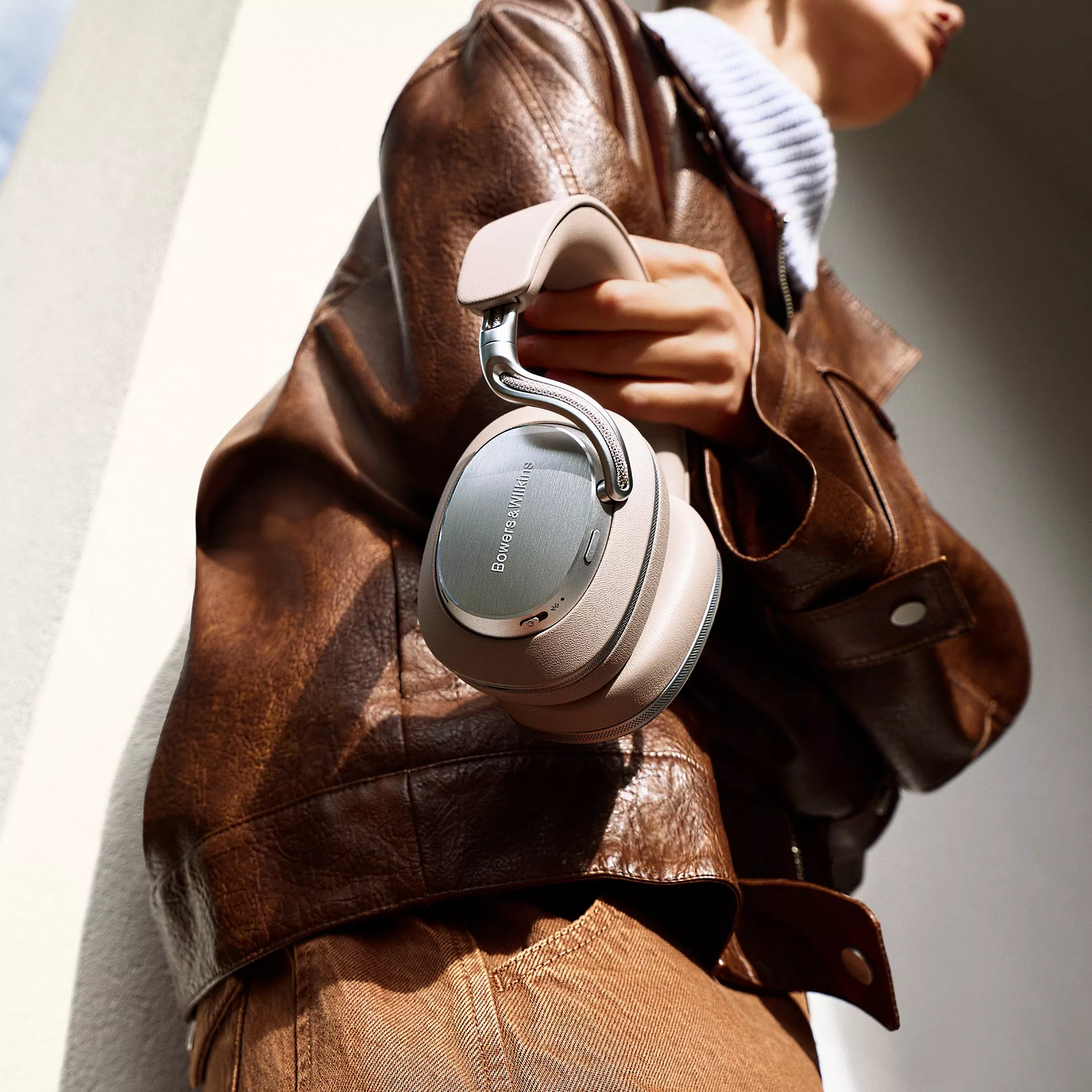 Person in brown leather jacket holding Bowers & Wilkins over-ear wireless noise-cancelling headphones outdoors
