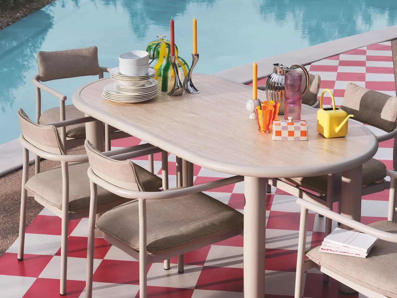 Outdoor patio dining table and cushioned chairs by a swimming pool, styled with plates, candles and colourful glassware