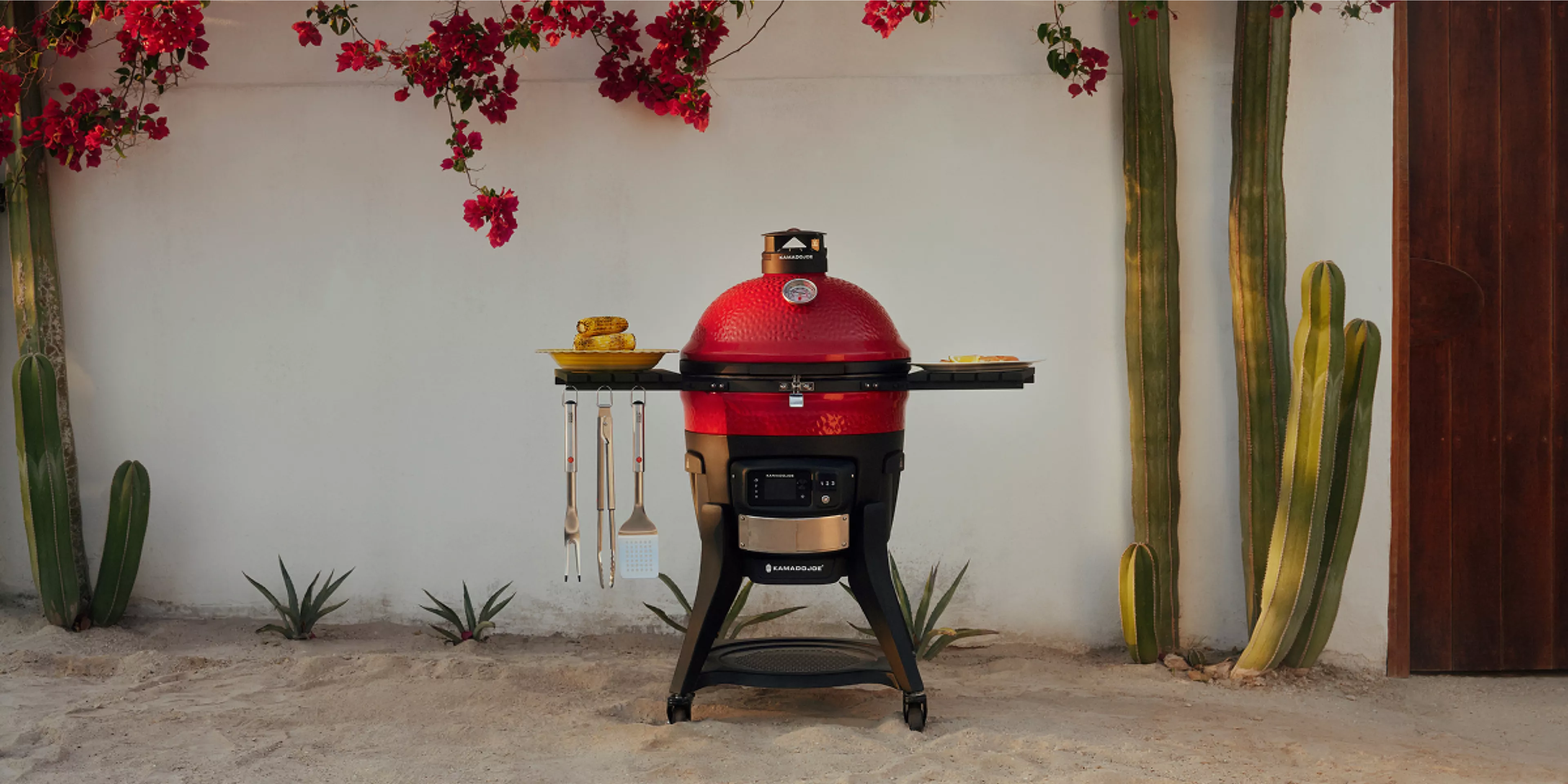 Red Kamado barbecue grill with side shelves displaying utensils and corn on the cob