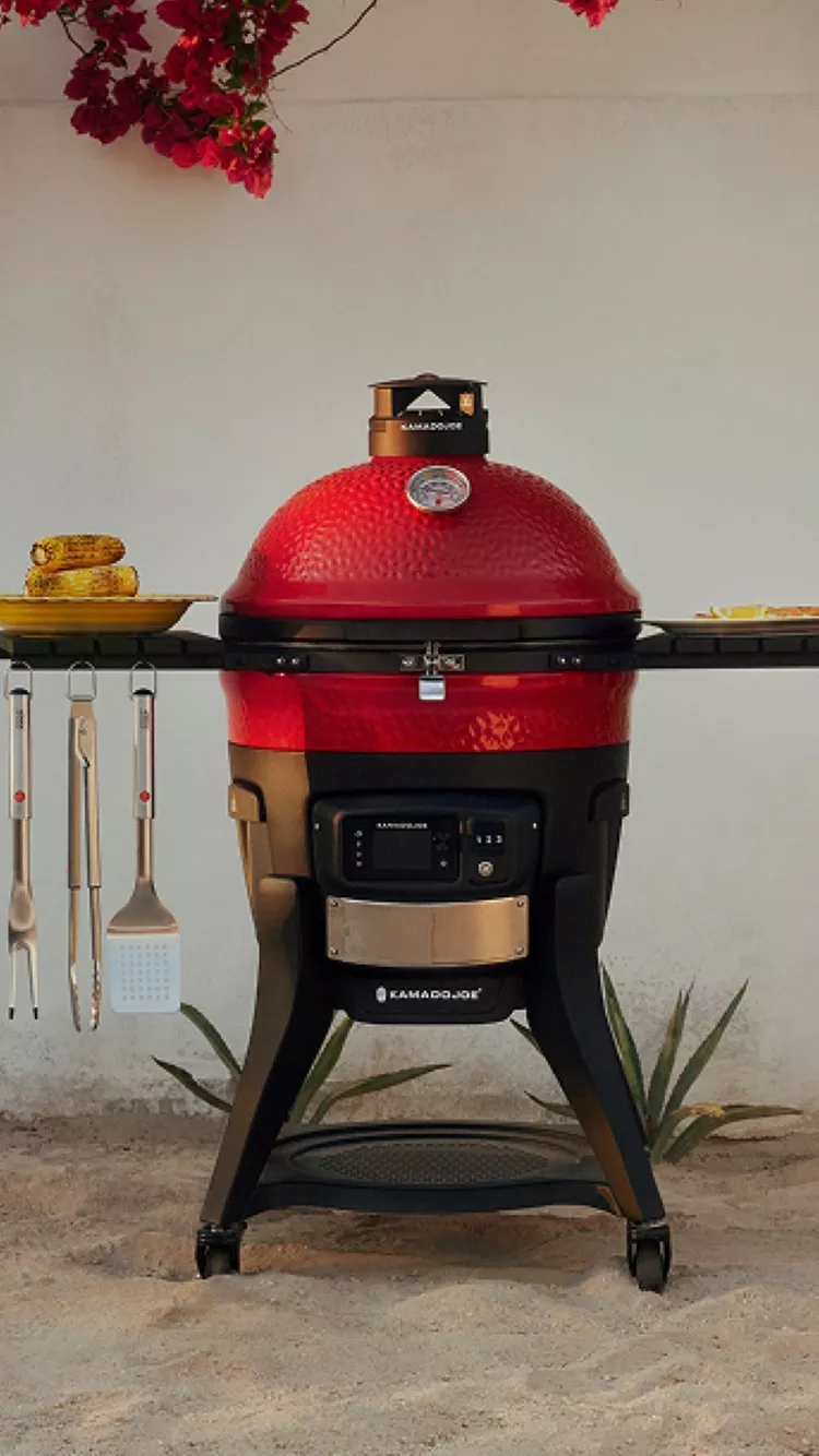Red Kamado barbecue grill with side shelves displaying utensils and corn on the cob