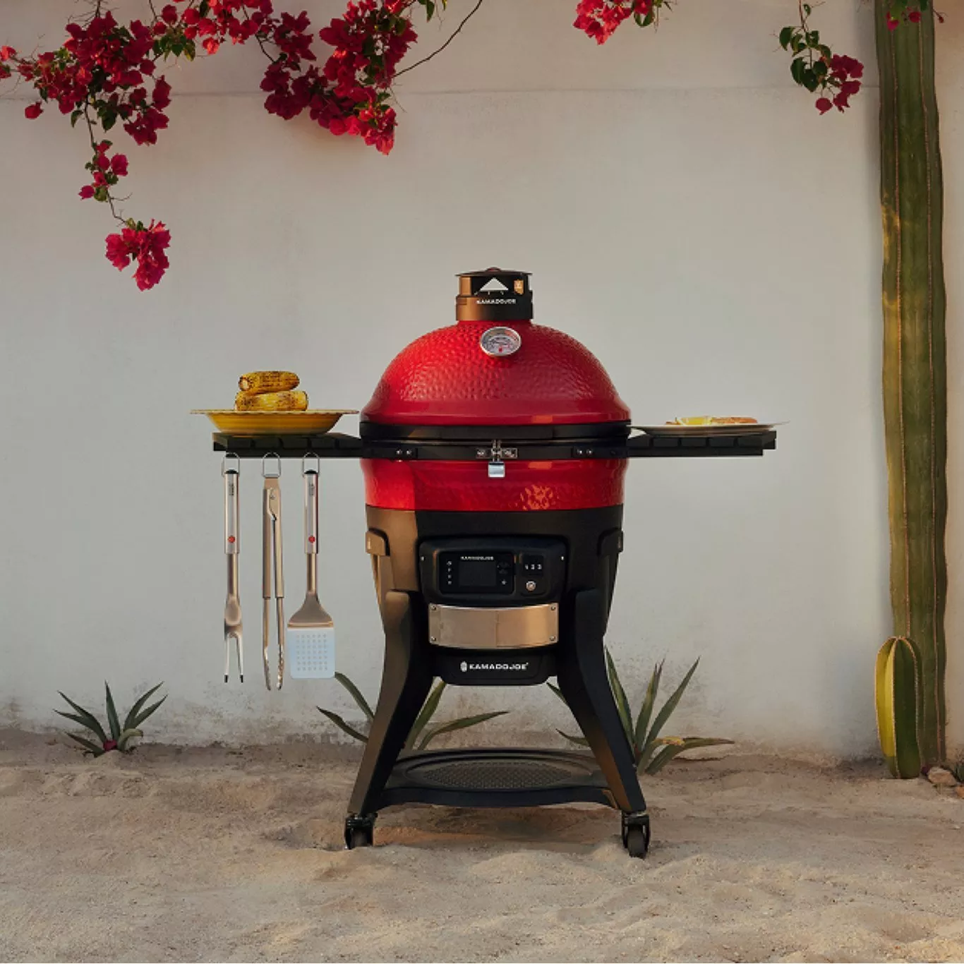 Red Kamado Joe ceramic barbecue grill with side shelves and hanging tools in an outdoor patio setting