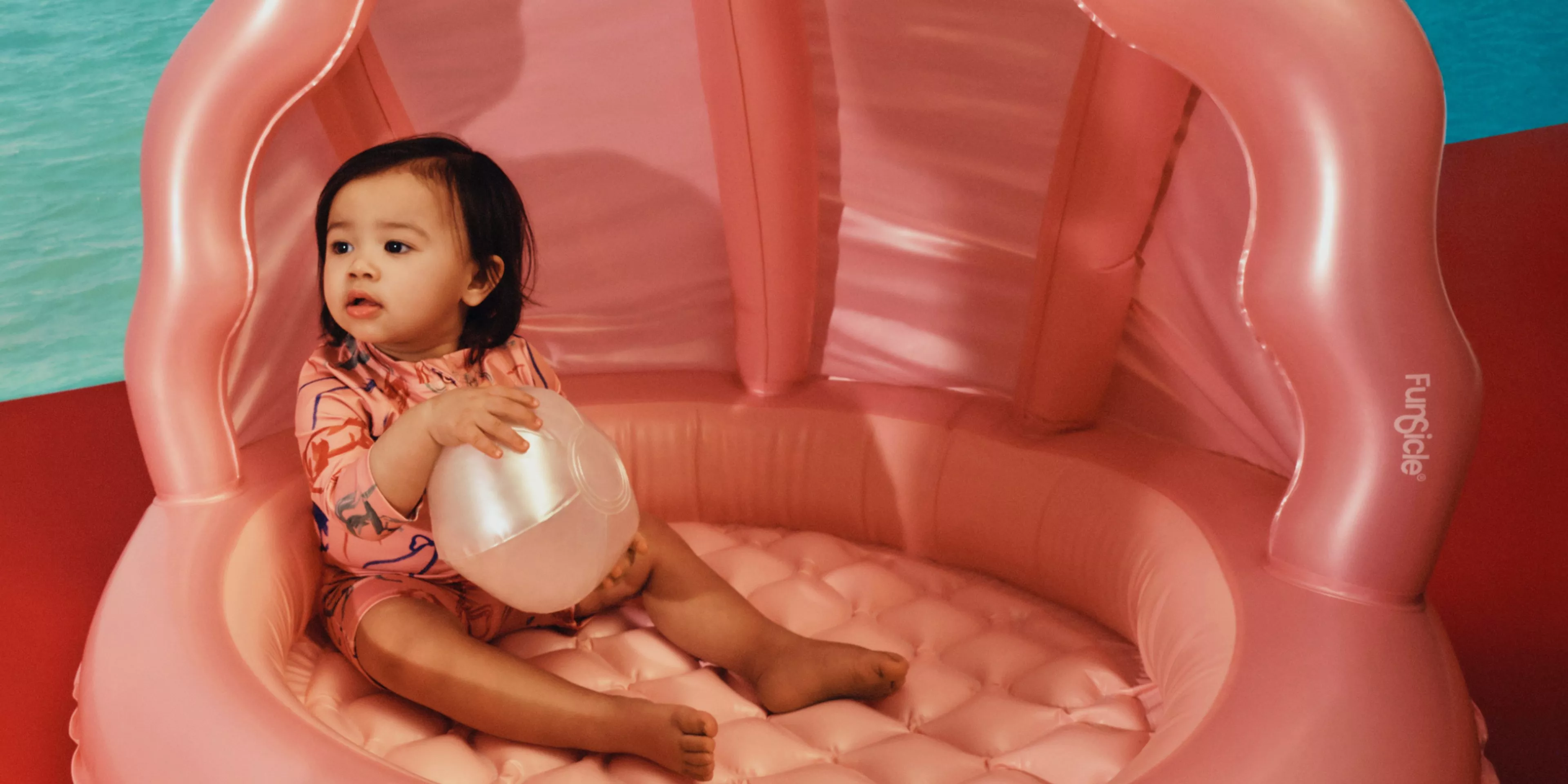 Toddler sitting in a pink inflatable shell pool holding a beach ball, with turquoise sea backdrop indoors