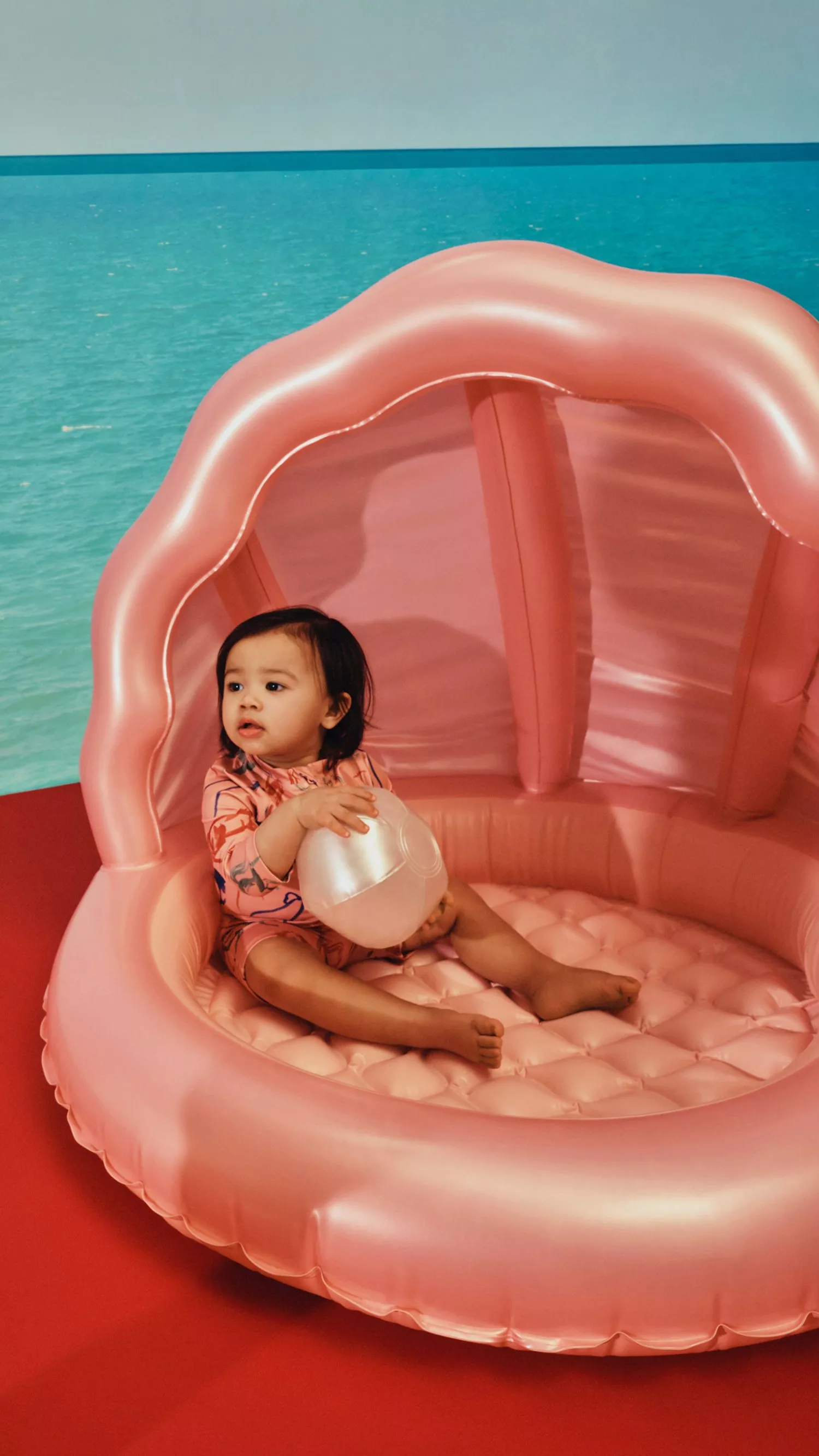 Toddler sitting in a pink inflatable shell pool holding a beach ball, with turquoise sea backdrop indoors