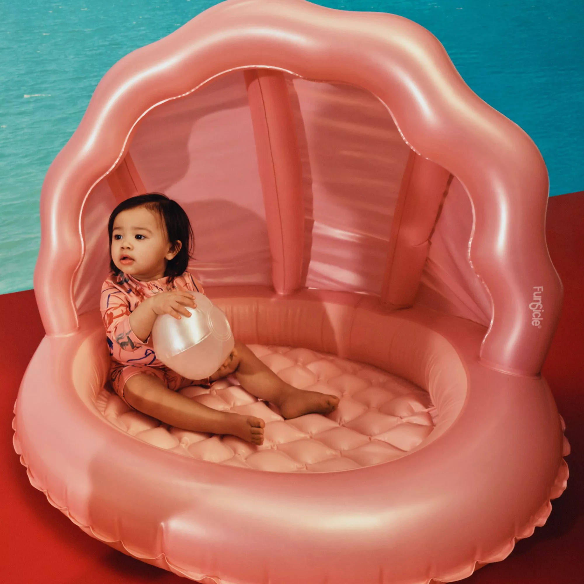 Toddler sitting in a pink inflatable shell paddling pool with canopy, holding a beach ball by the water