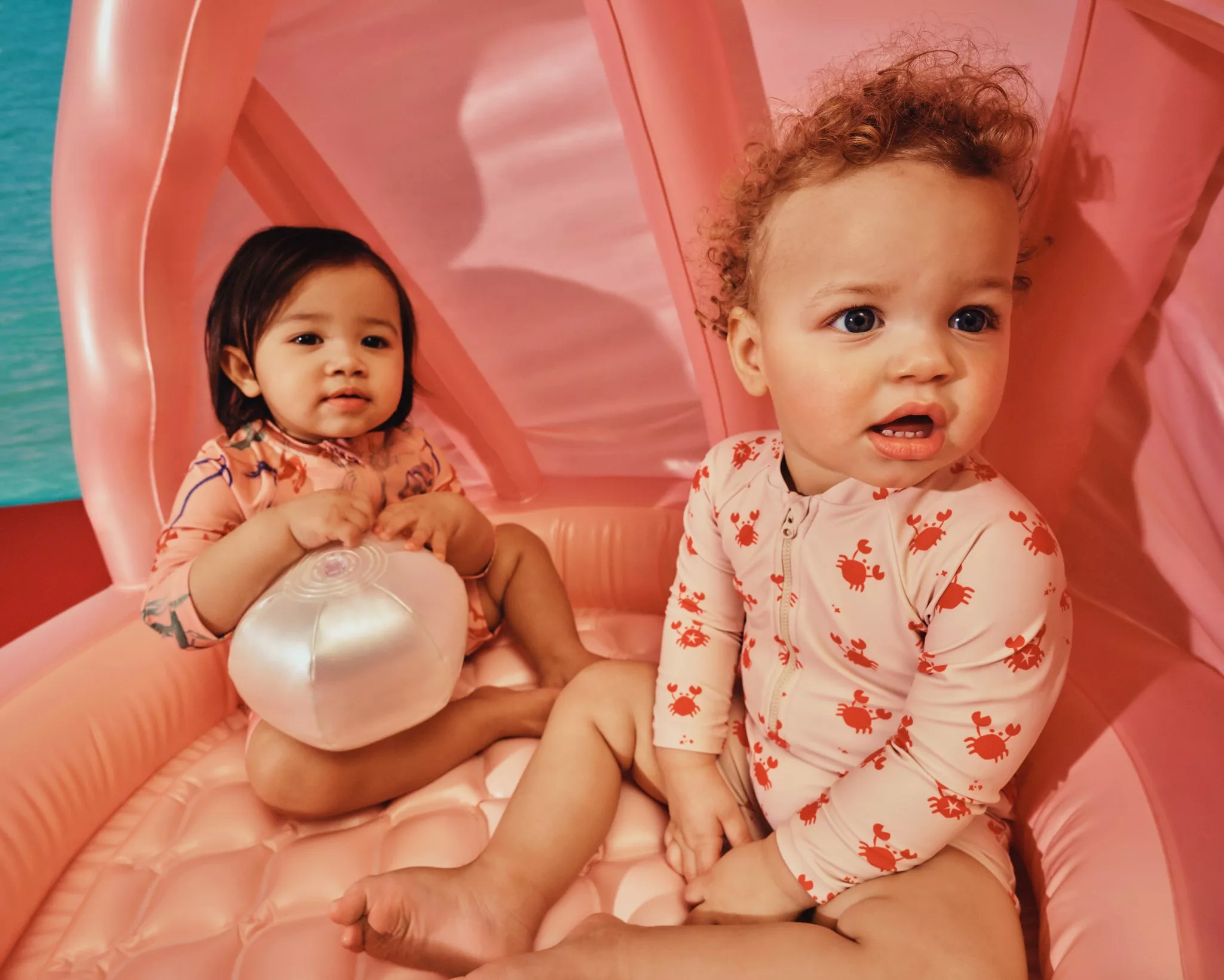 Two toddlers in crab-print swimwear sitting on a pink inflatable pool float with a beach ball, summer fun scene