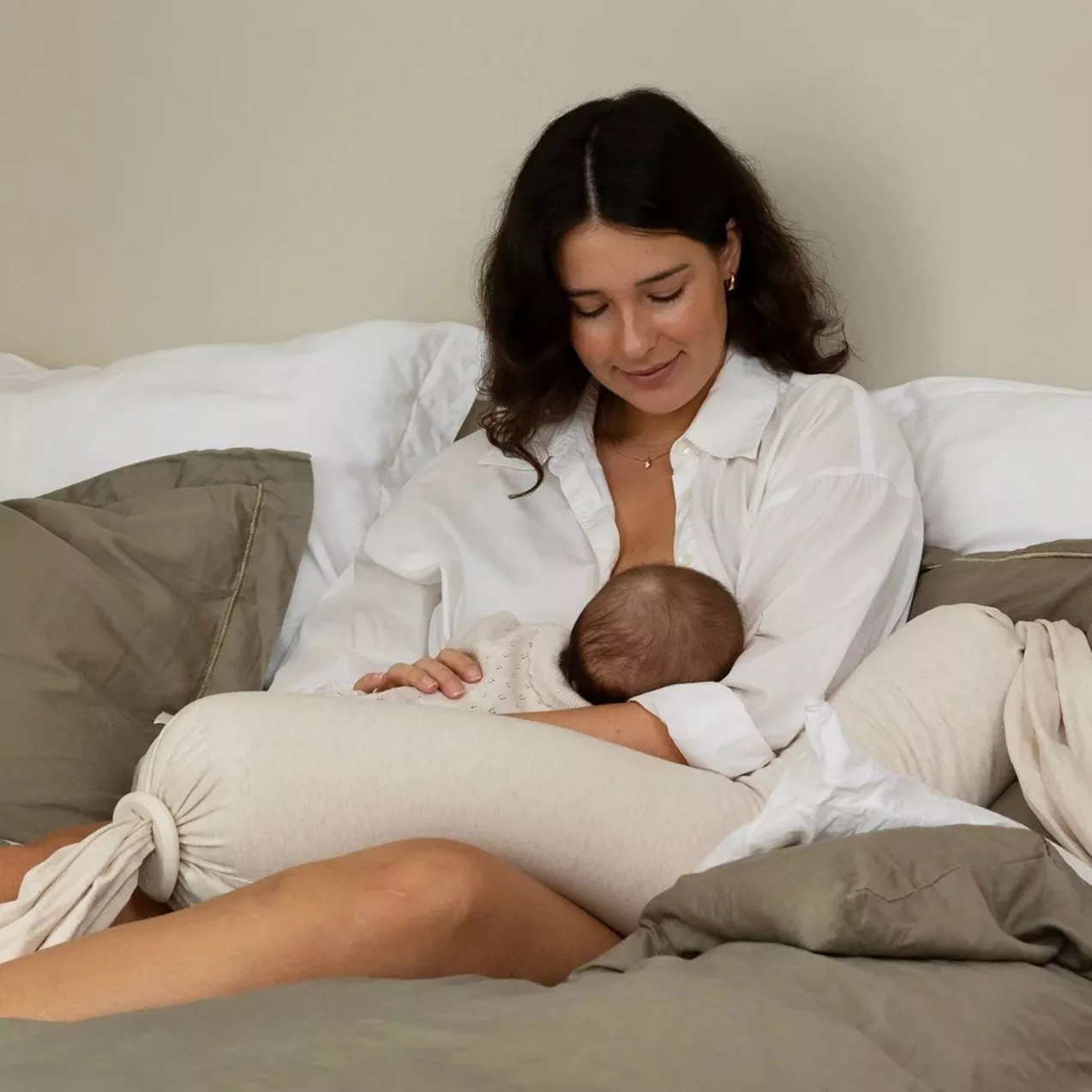 Mother breastfeeding baby on bed with pillows, cosy neutral bedroom scene, gentle bonding and newborn care