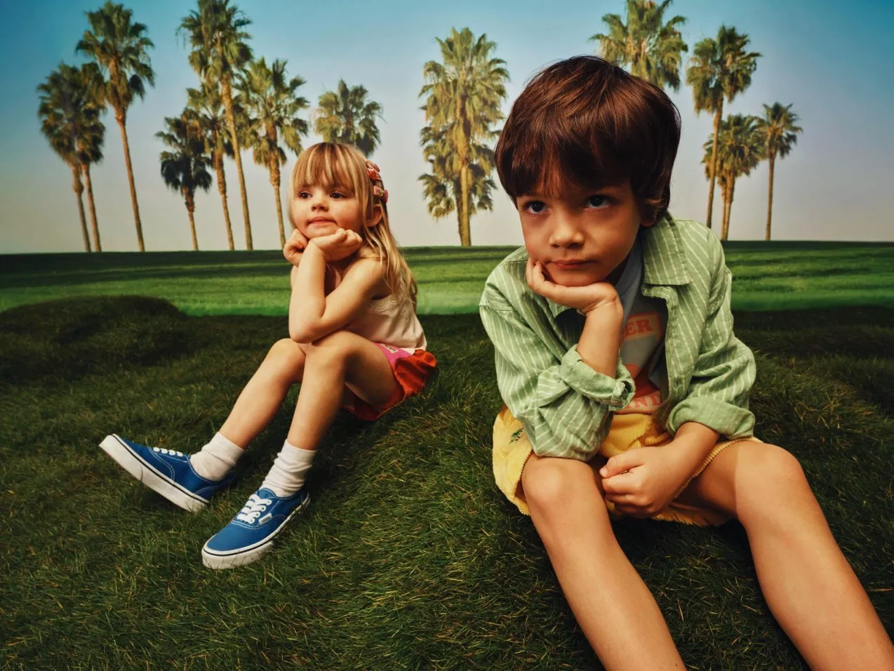 Two young children sitting on a grassy park lawn with palm trees, resting chin on hands, sunny day.