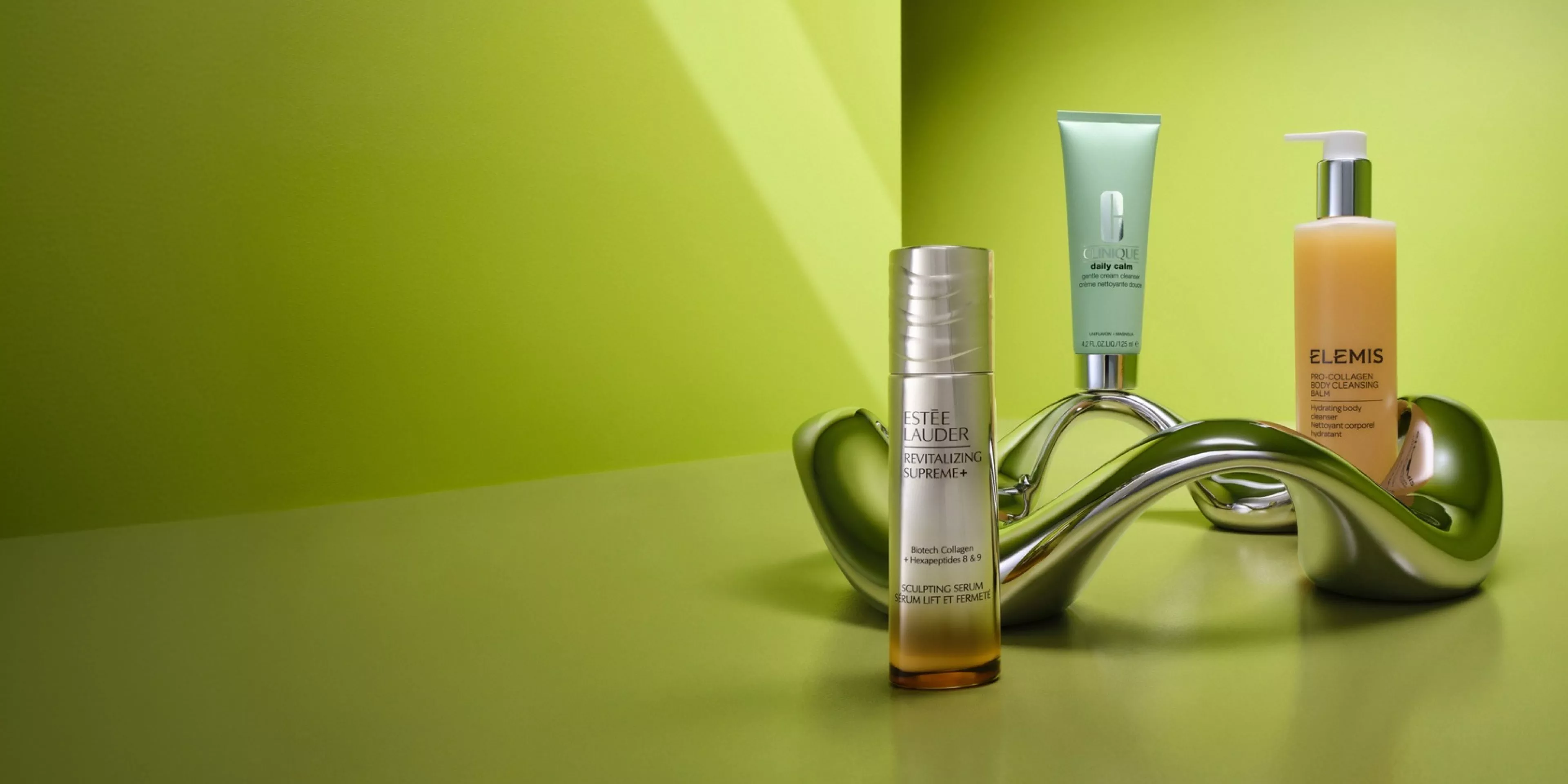 Estée Lauder serum, Clinique cream and Elemis body cleanser displayed on green background with metallic sculpture