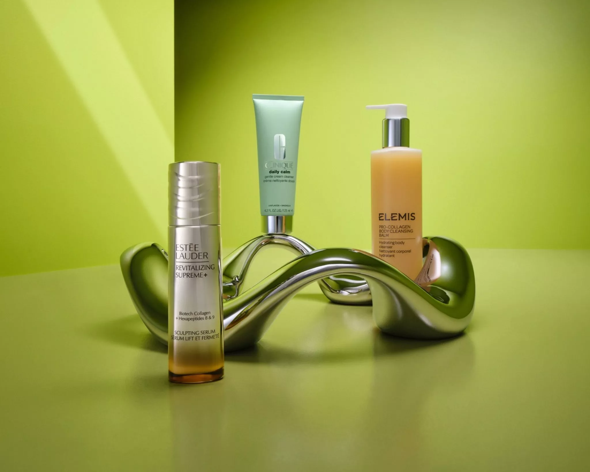 Luxury skincare products on green backdrop, including Estée Lauder serum, Clinique cleanser and Elemis body balm