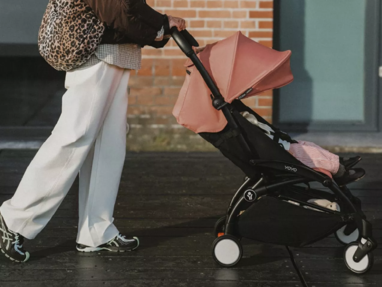 Parent pushing compact pink and black baby stroller pushchair on pavement outside brick building